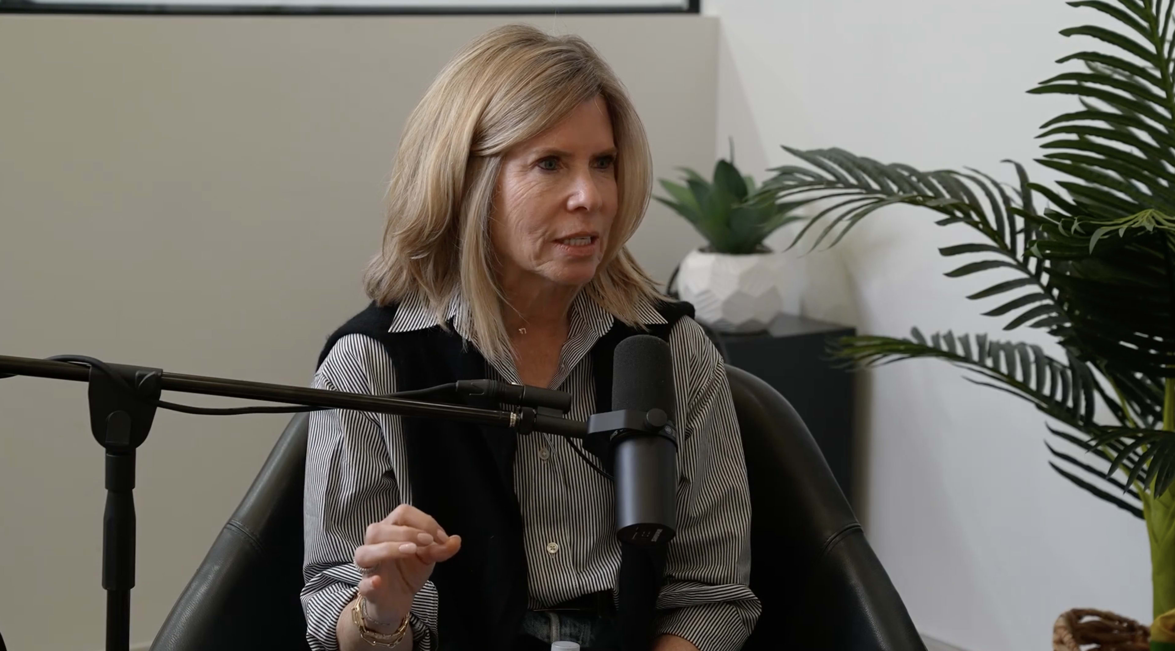 A woman sits in a chair, speaking into a microphone during a podcast recording, with plants visible in the background.