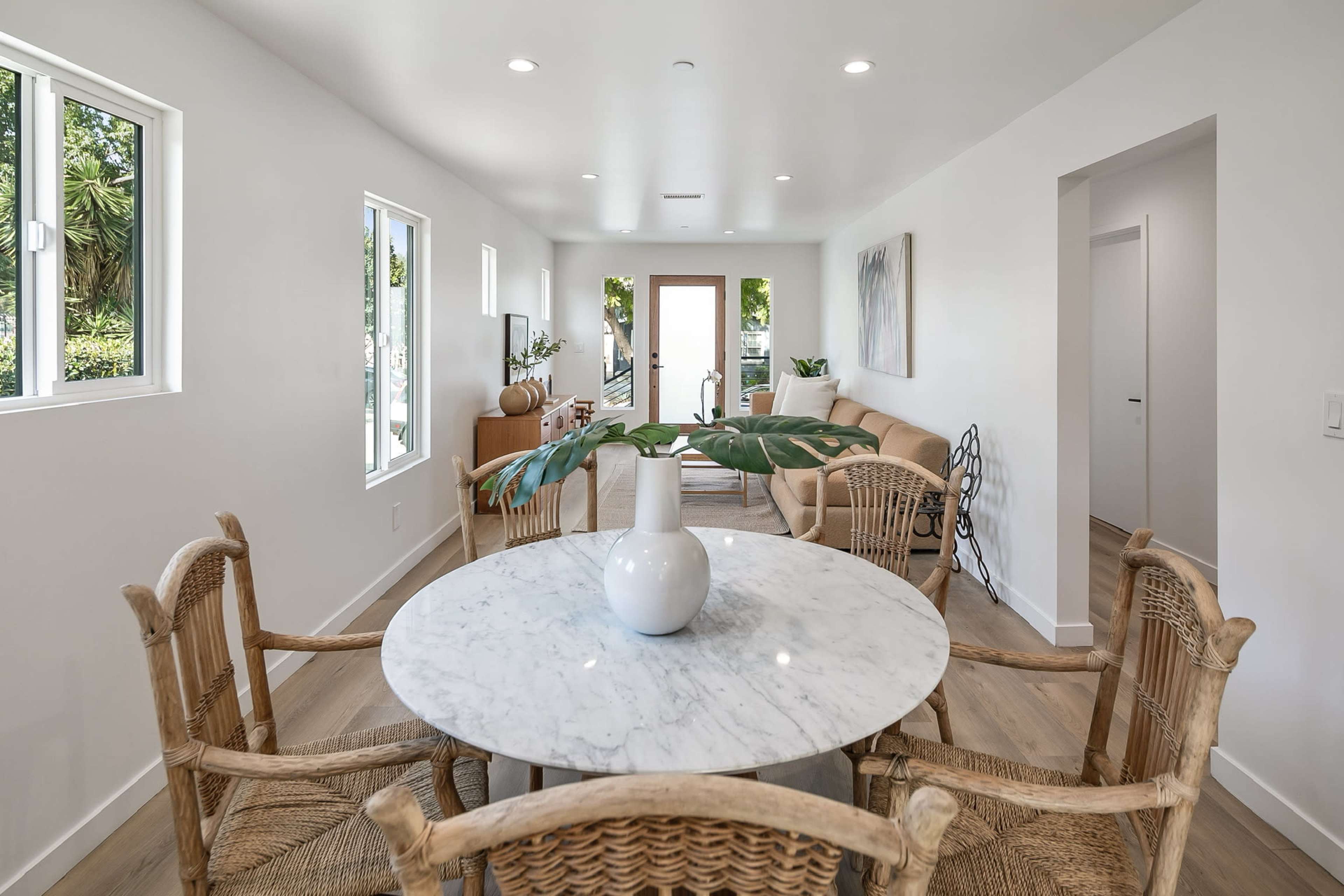 A round marble-topped dining table is surrounded by four wicker chairs in a bright, modern living space with large windows and an open floor plan.