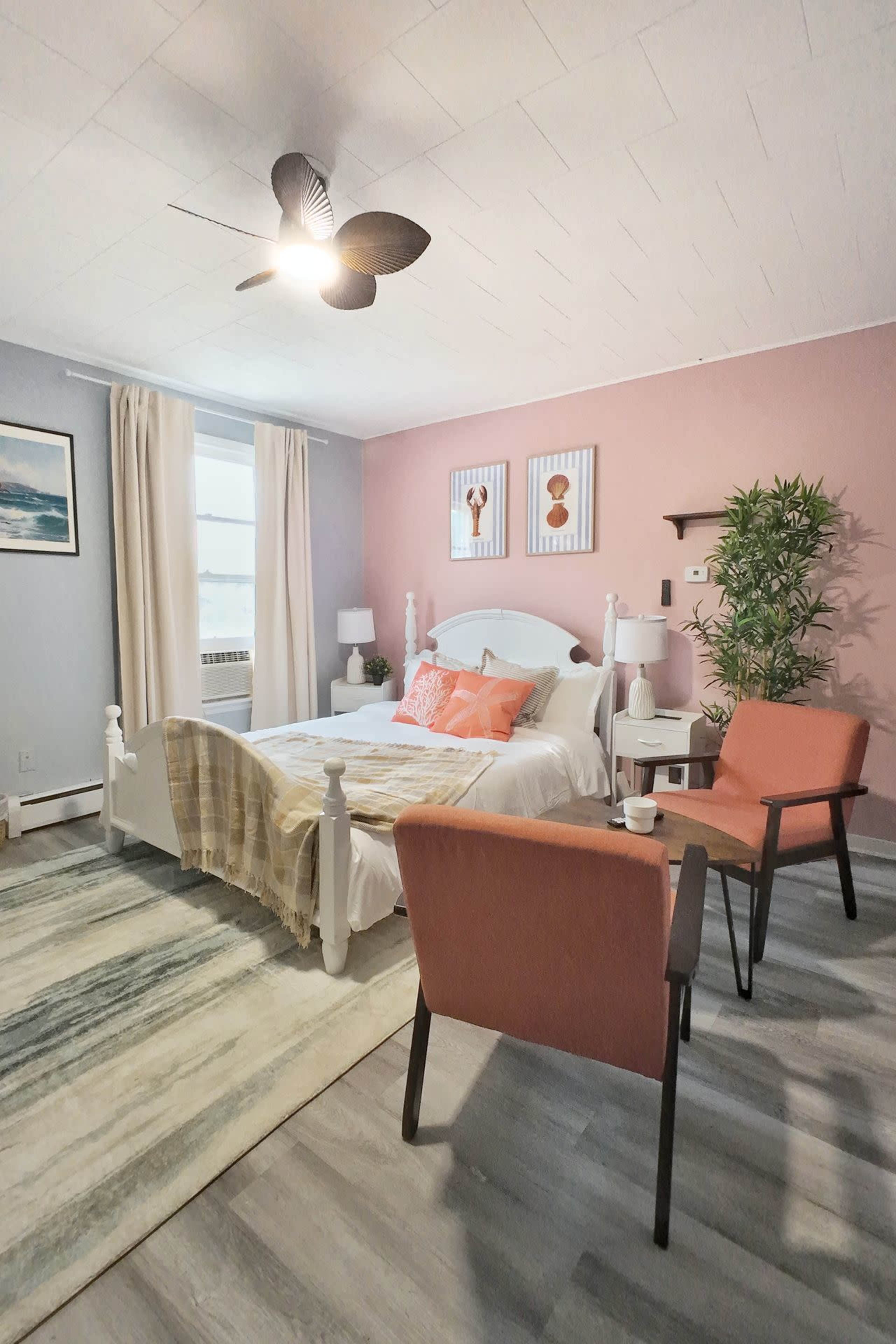 The image shows a well-decorated bedroom featuring a bed with colorful pillows, two chairs, a rug, and framed artwork on the walls.