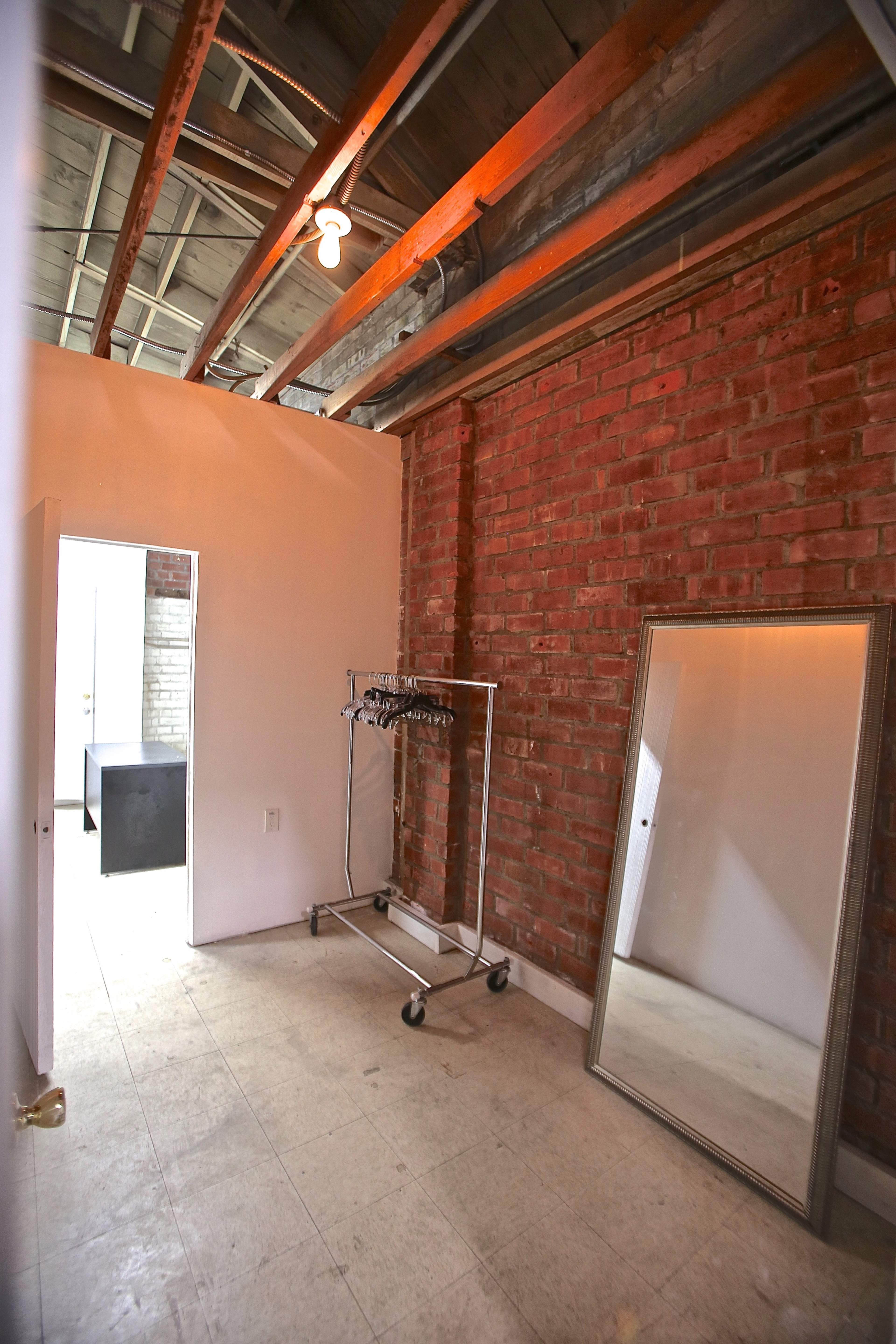 The image shows a small interior space with exposed brick walls, a full-length mirror, and a clothing rack mounted on wheels.