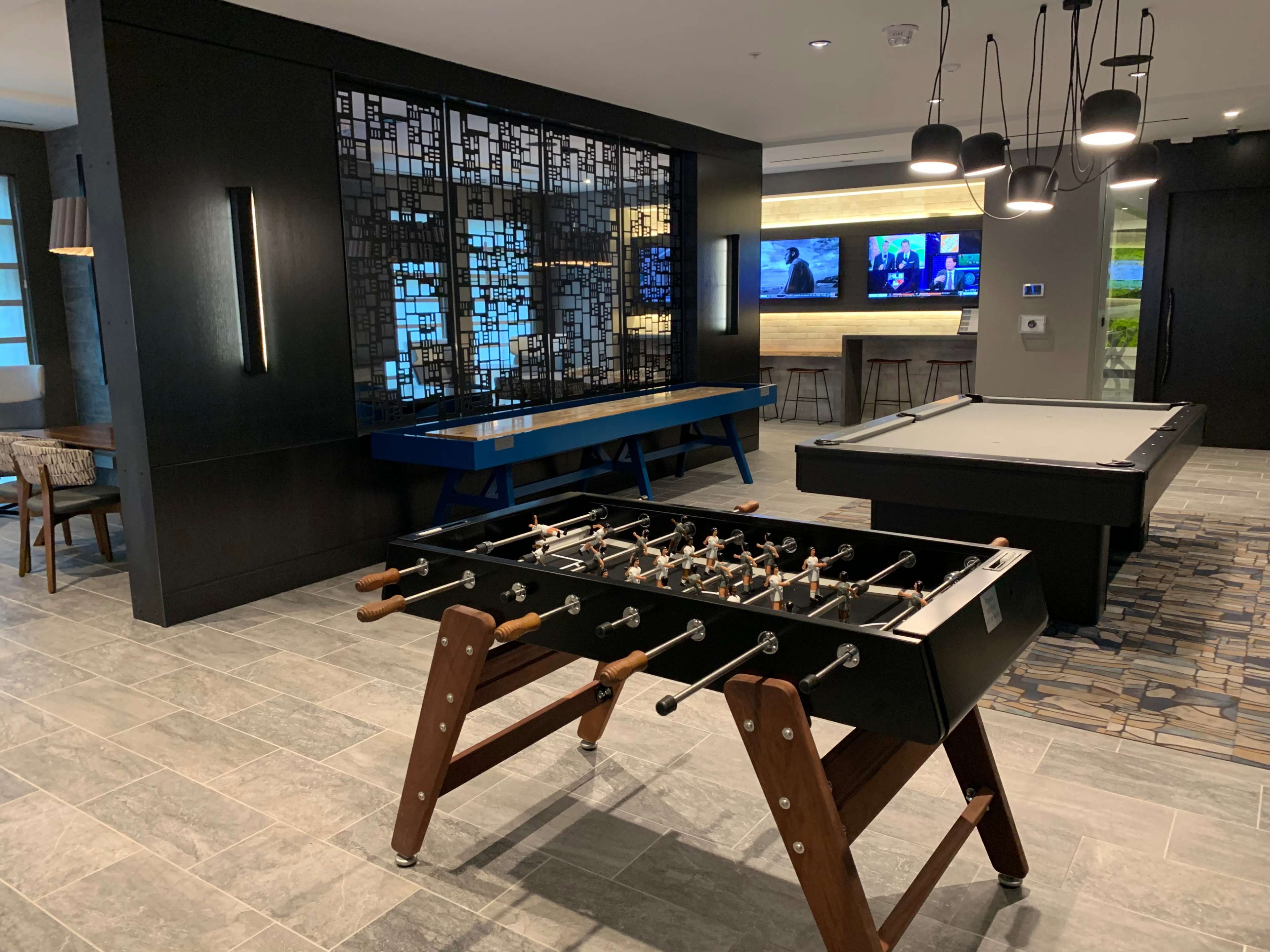 A recreational room featuring a foosball table in the foreground, a pool table in the background, and several screens mounted on the wall.