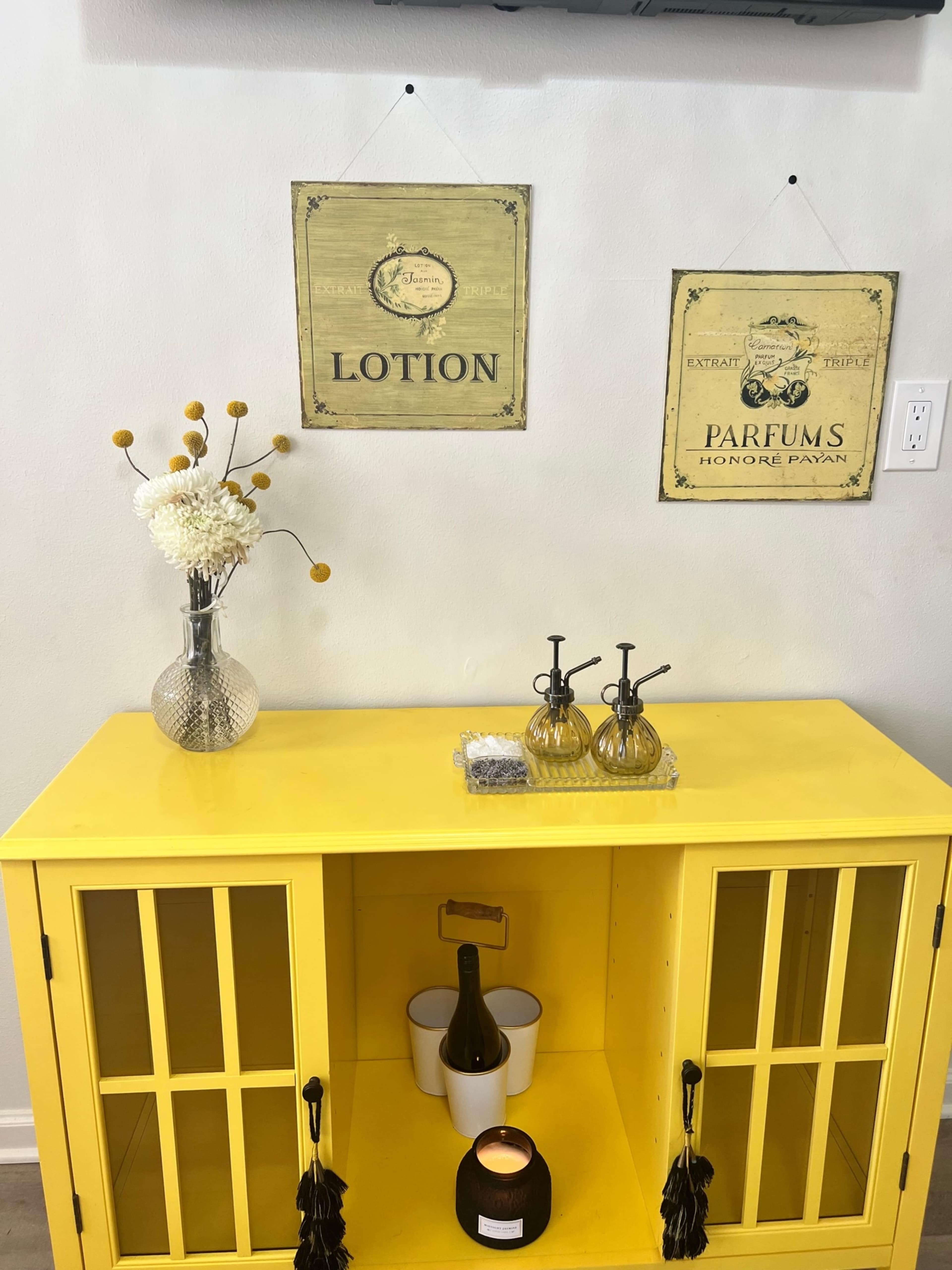 A yellow cabinet displays two framed signs labeled "LOTION" and "PARFUMS," alongside a vase of flowers, a candle, a bottle, and two glass dispensers.