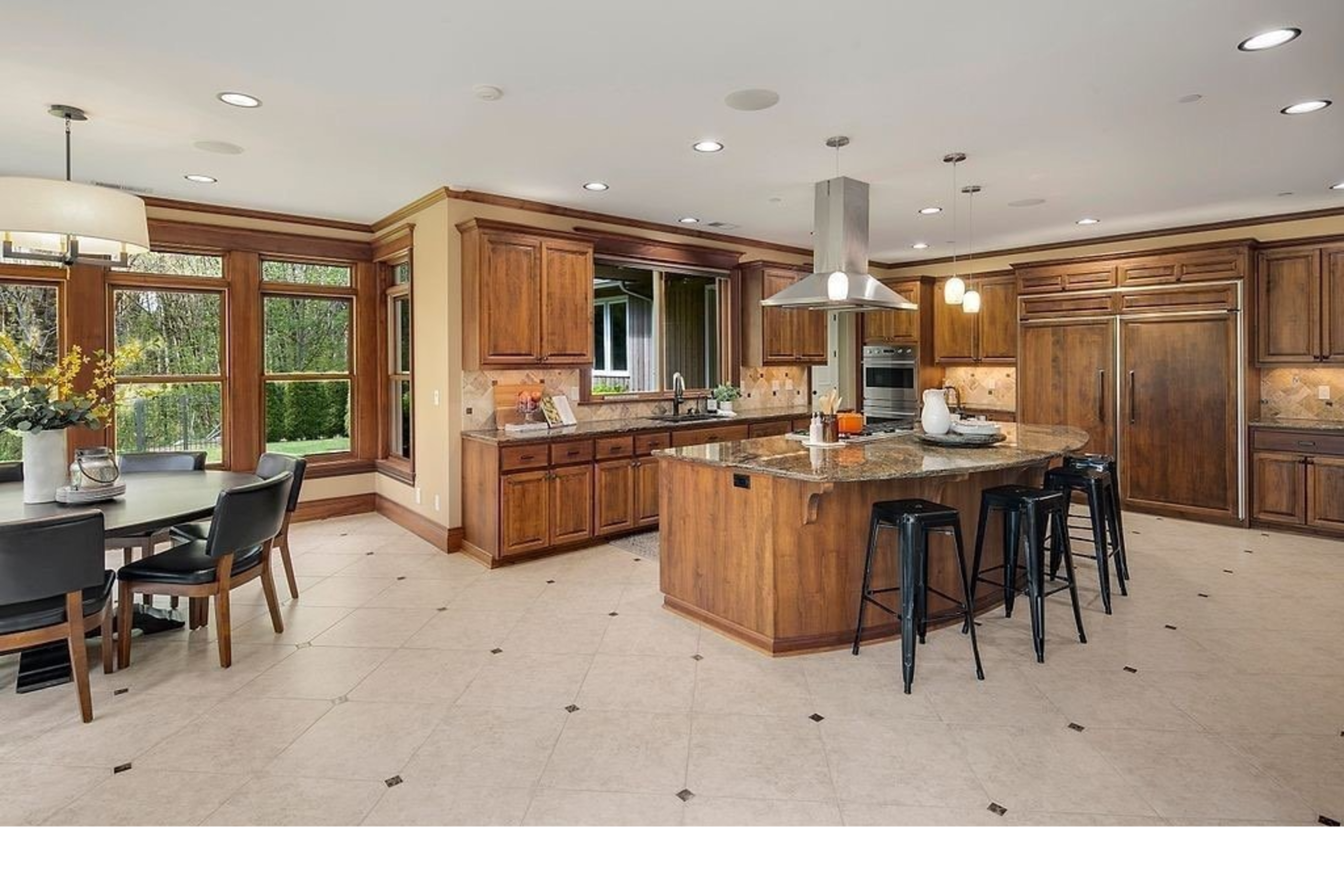The image shows a spacious kitchen with wooden cabinetry, a large central island, and a dining area set with a circular table and black chairs.