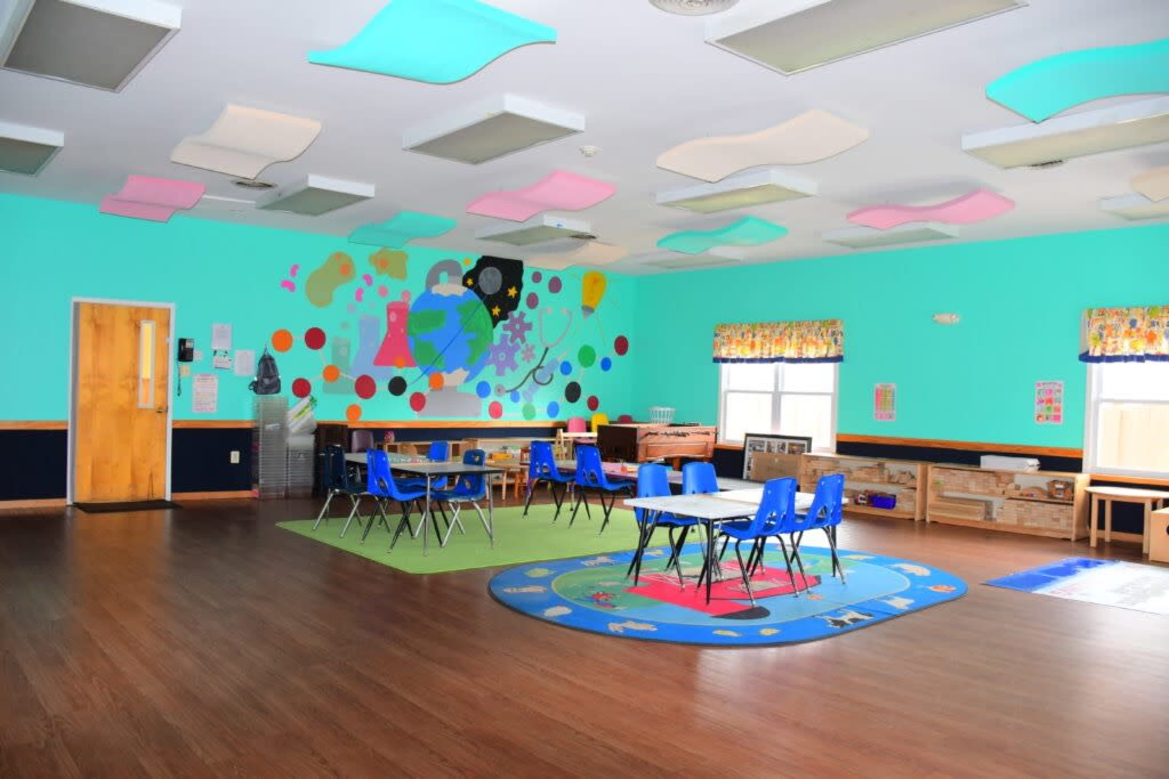 A brightly colored classroom features blue walls, multiple rugs, and several tables and chairs arranged for children's activities.