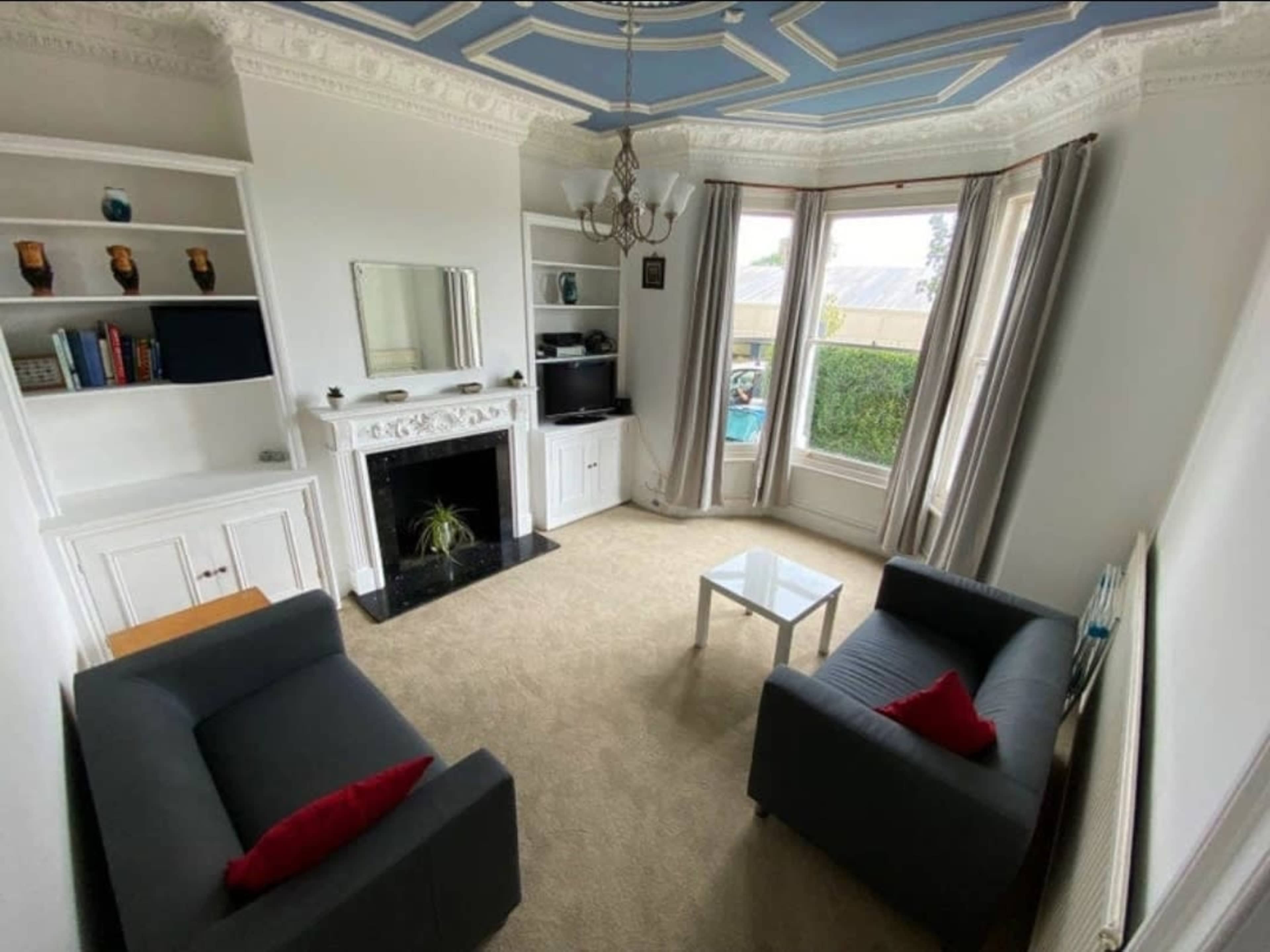A living room features two gray sofas with red cushions, a glass coffee table, a fireplace, built-in shelves, and large windows overlooking a garden.
