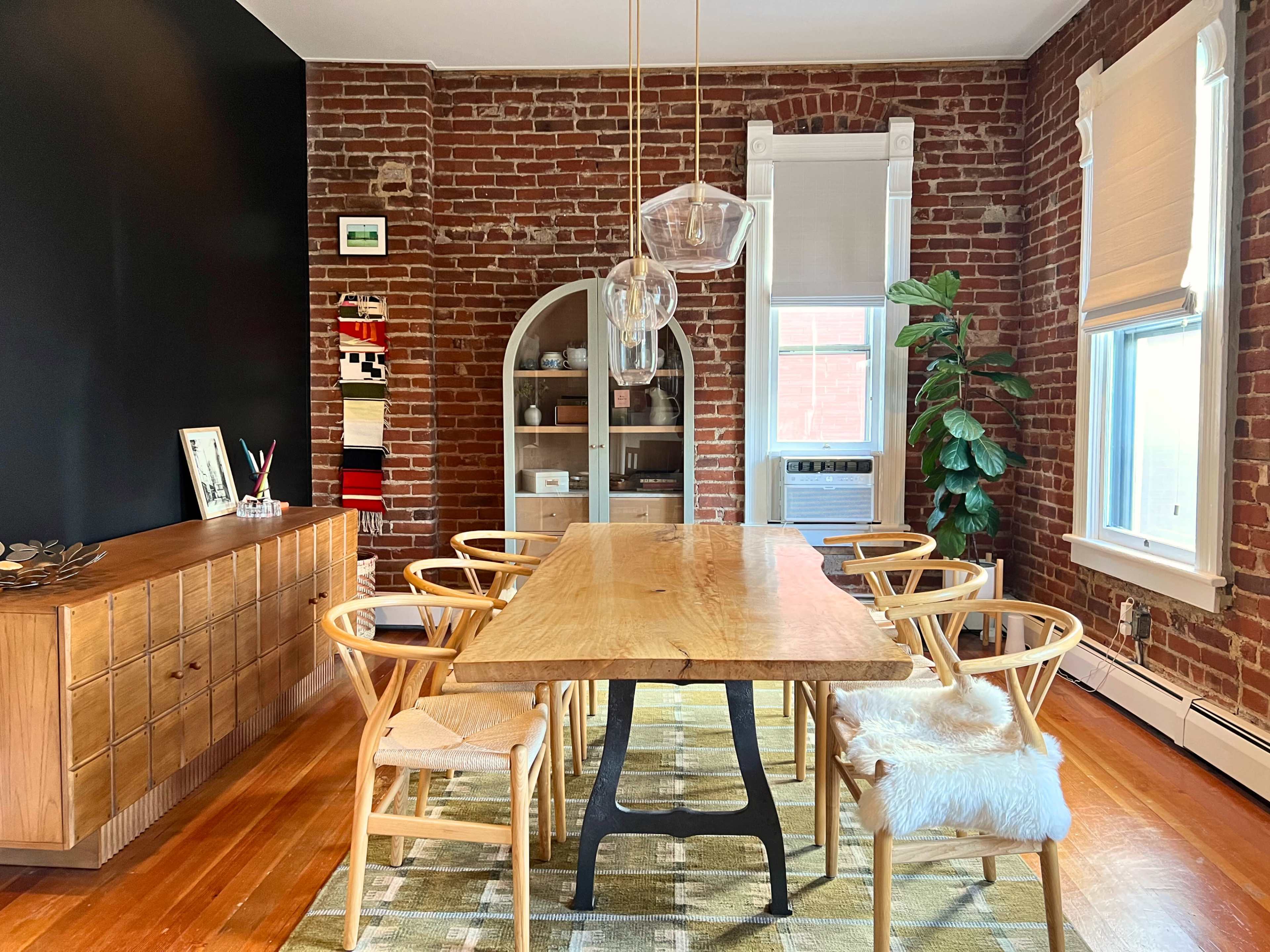 The image shows a modern dining room featuring a long wooden table surrounded by six chairs, set against a brick wall with a shelving unit and a large plant in the corner.