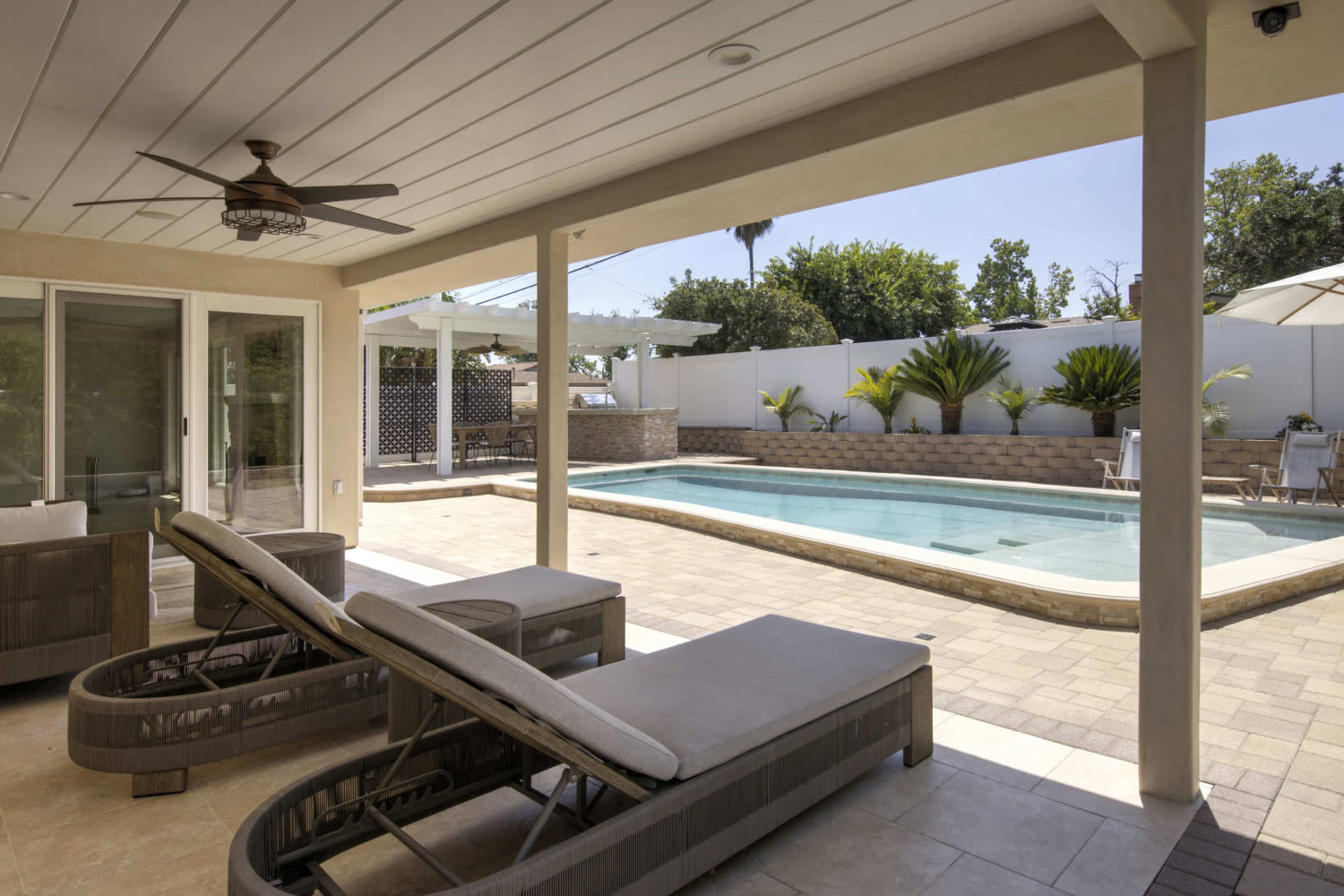 The image shows a modern outdoor patio area with two lounge chairs overlooking a swimming pool surrounded by landscaped greenery.
