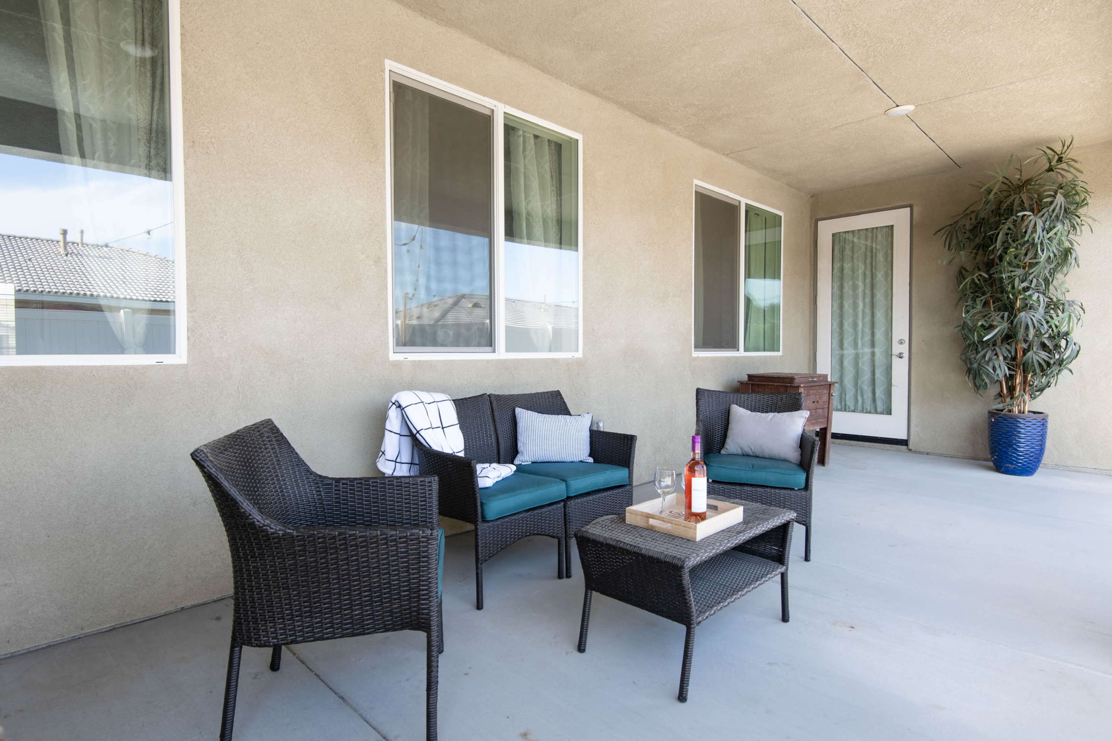 The image shows a patio area with four wicker chairs, a small table, and a large potted plant, all situated against a beige wall and large windows.