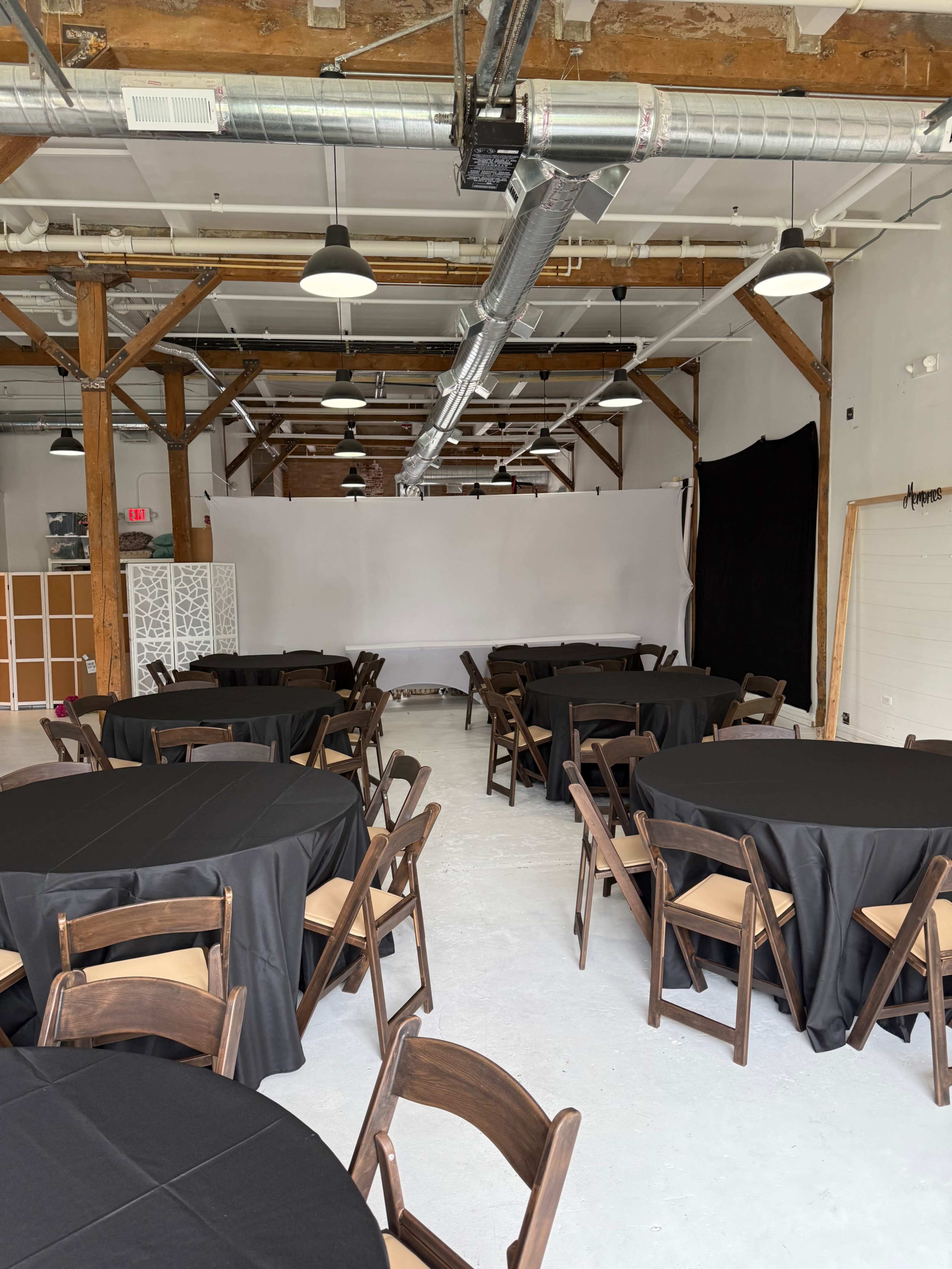 The image shows a spacious indoor venue with wooden beams, several round tables covered in black tablecloths, and a plain white backdrop at one end.