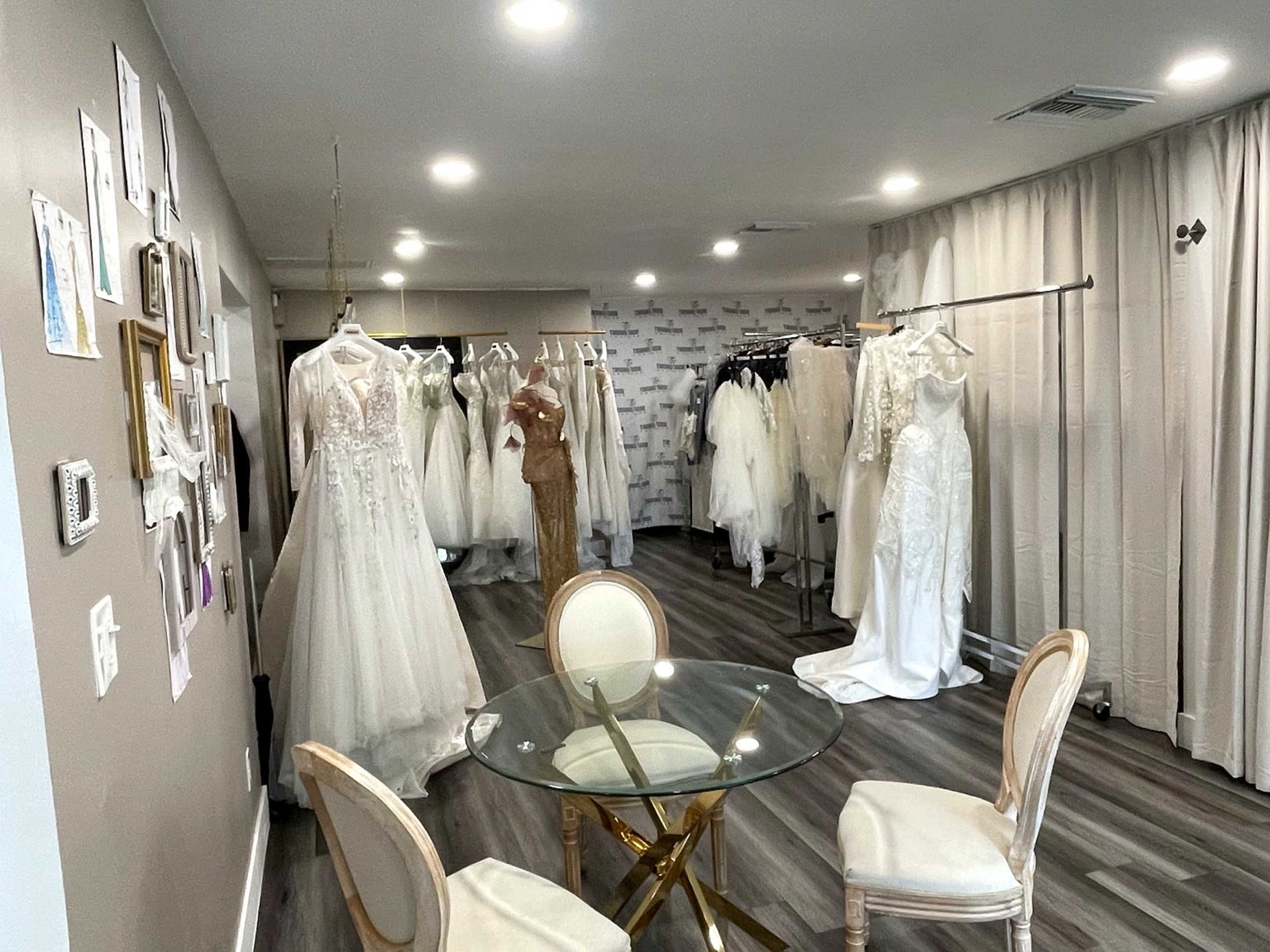 The image depicts a bridal boutique featuring several wedding dresses displayed on racks and hangers, with a small round table and chairs in the center of the room.