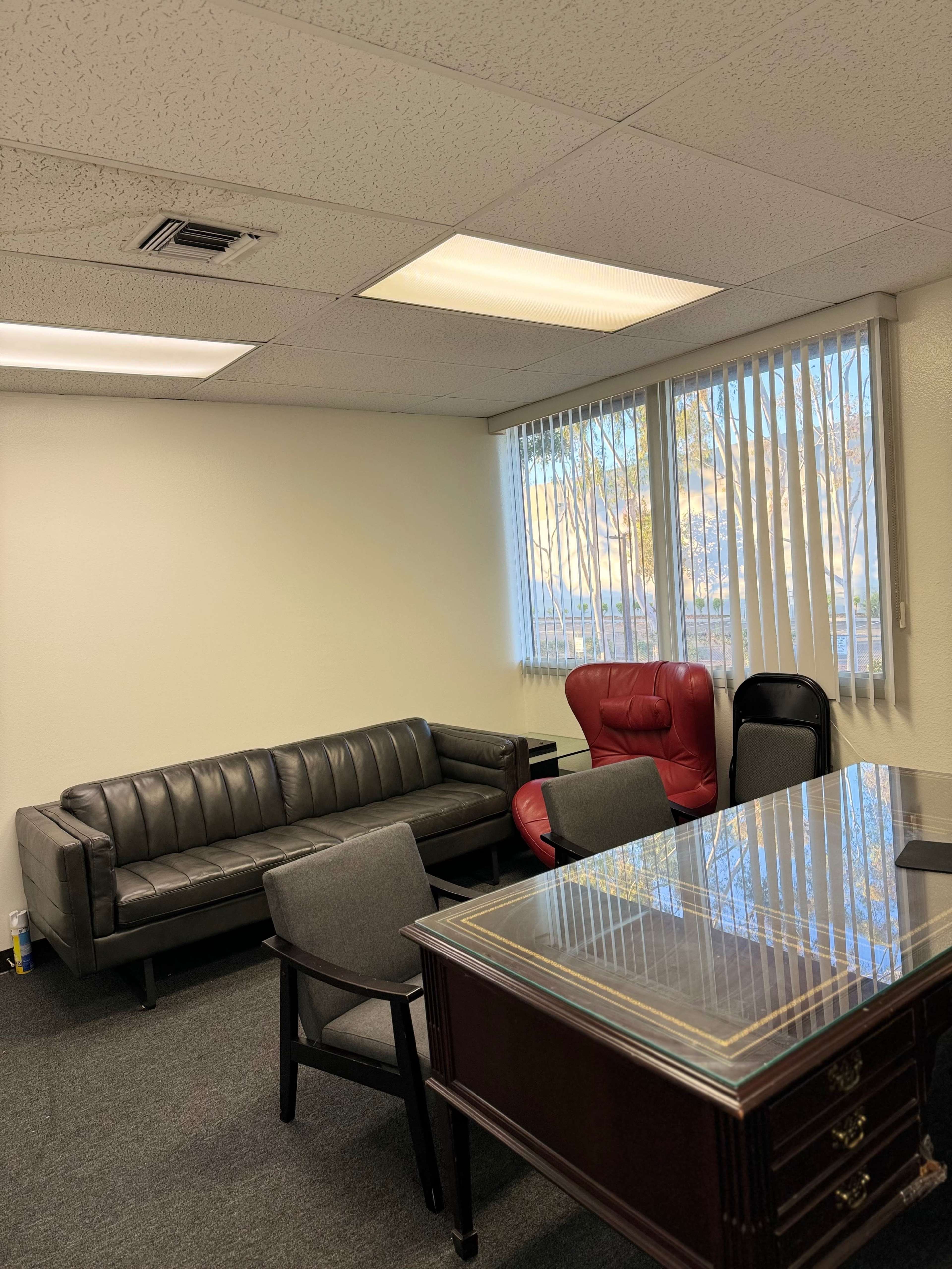 The image shows an office space with a black sofa, a red chair, several gray chairs, and a wooden desk near a window with blinds.