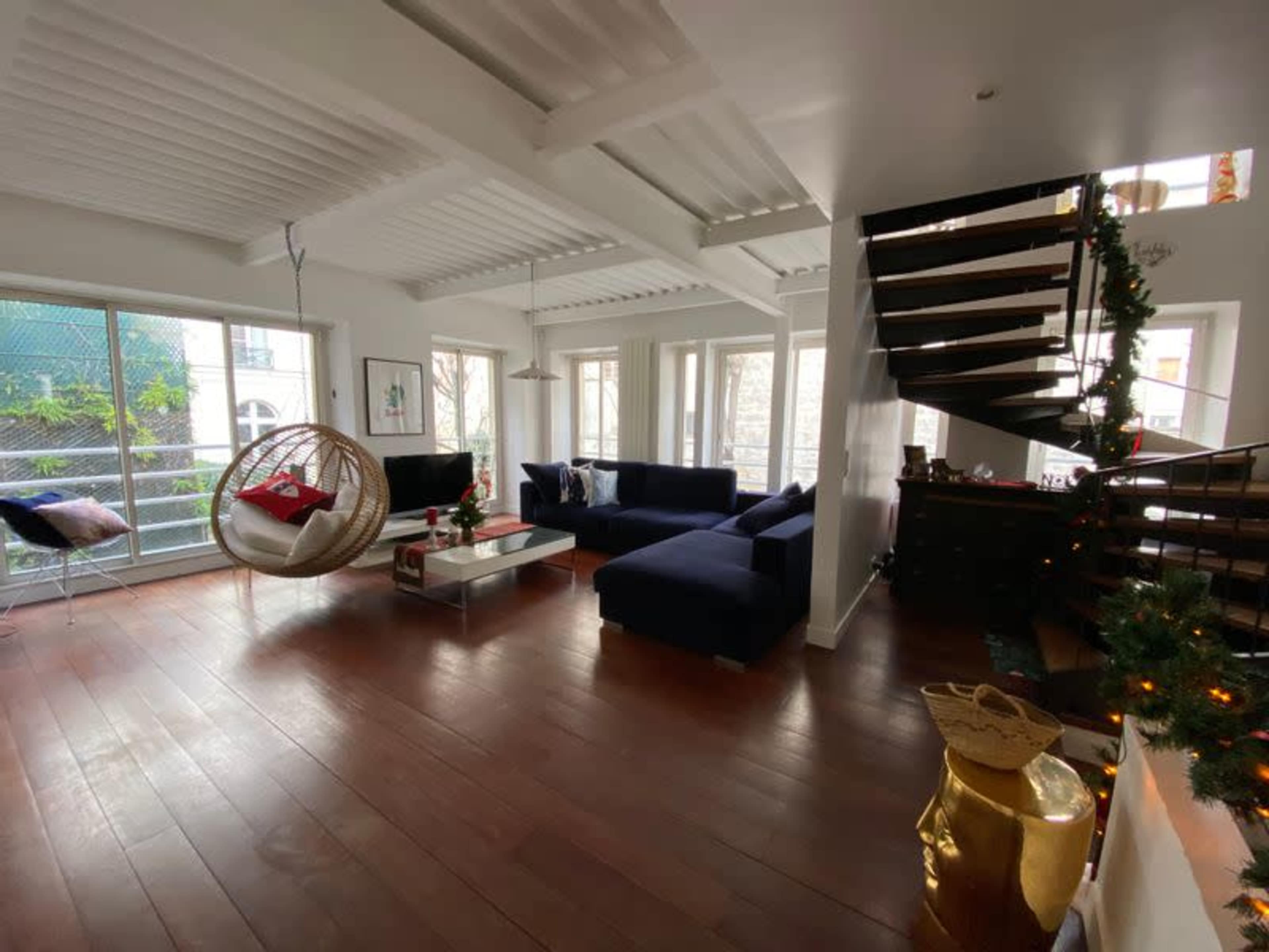 A modern living room features a large sectional couch, a hanging chair, and a staircase with wooden steps, surrounded by large windows that provide natural light.