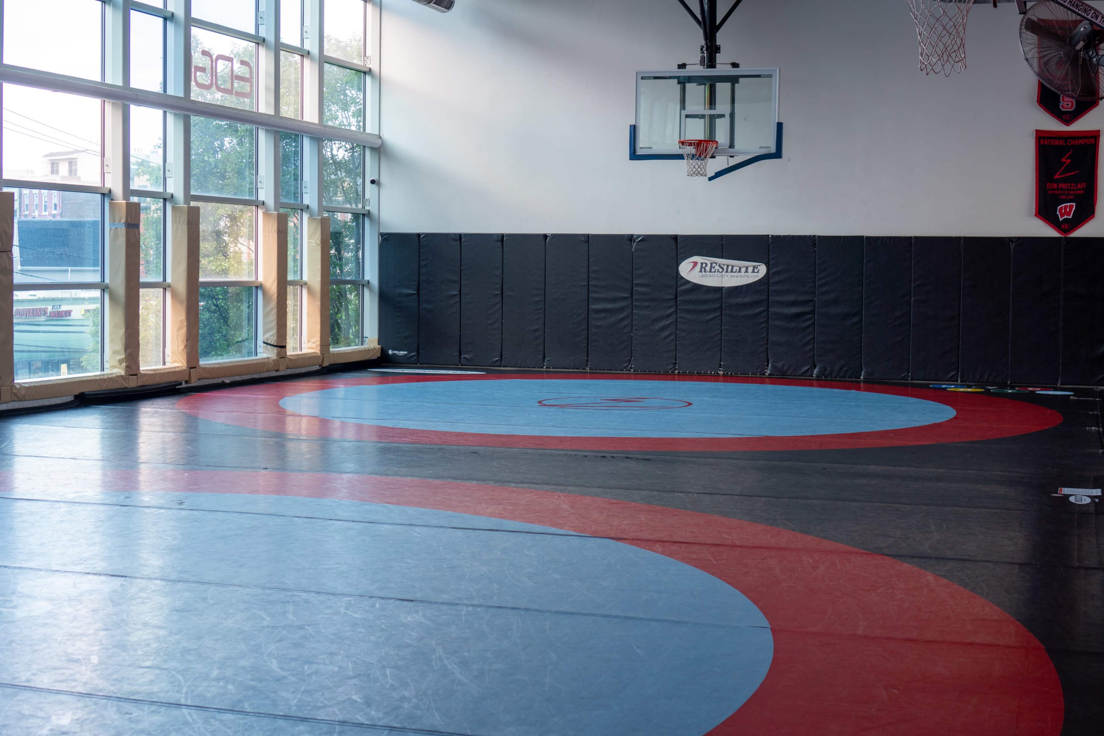 A gymnasium with a wrestling mat featuring blue and red circles and a basketball hoop in the background.