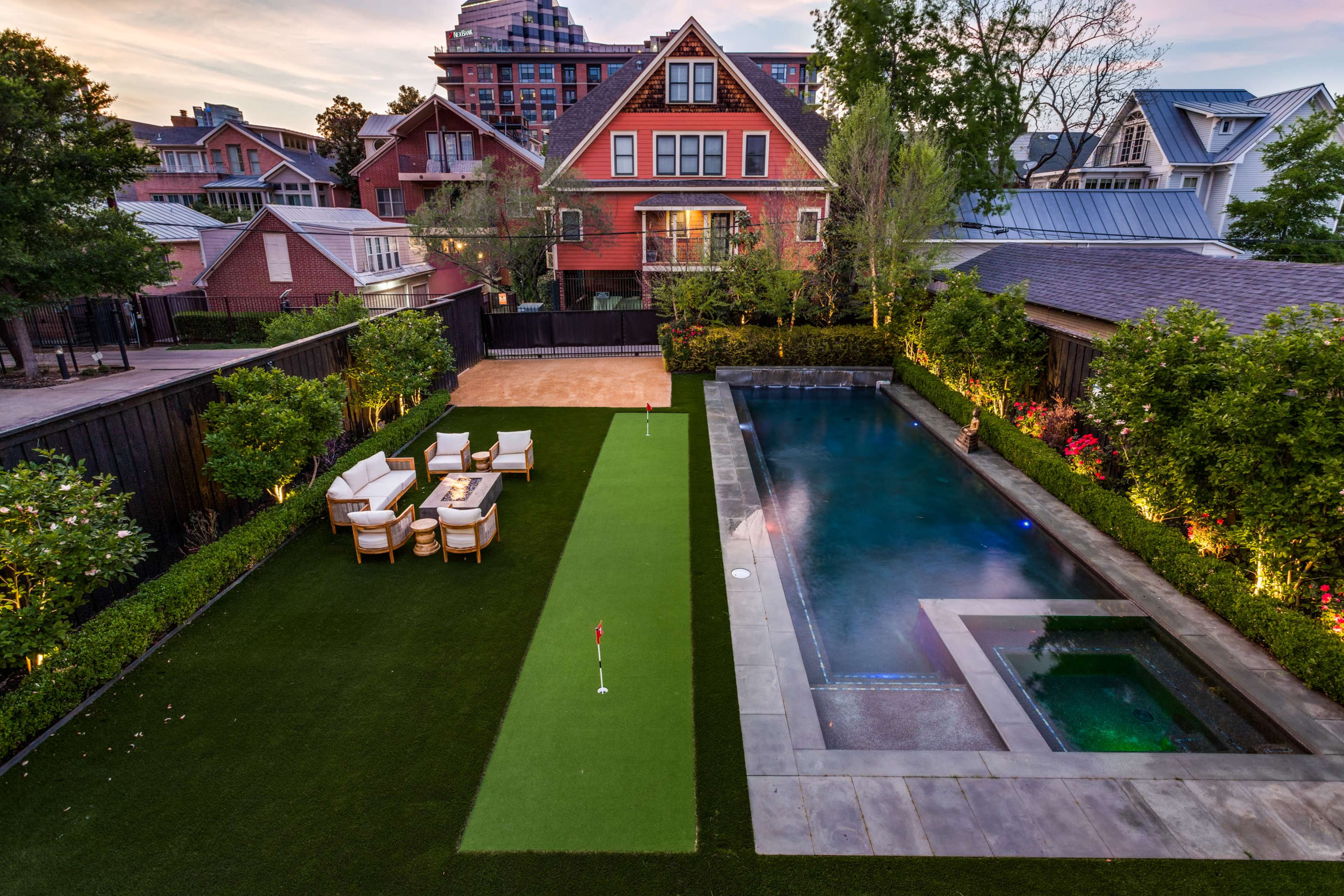 A well-maintained backyard featuring a green putting green and a rectangular swimming pool, bordered by manicured shrubs and a red-brick house in the background.