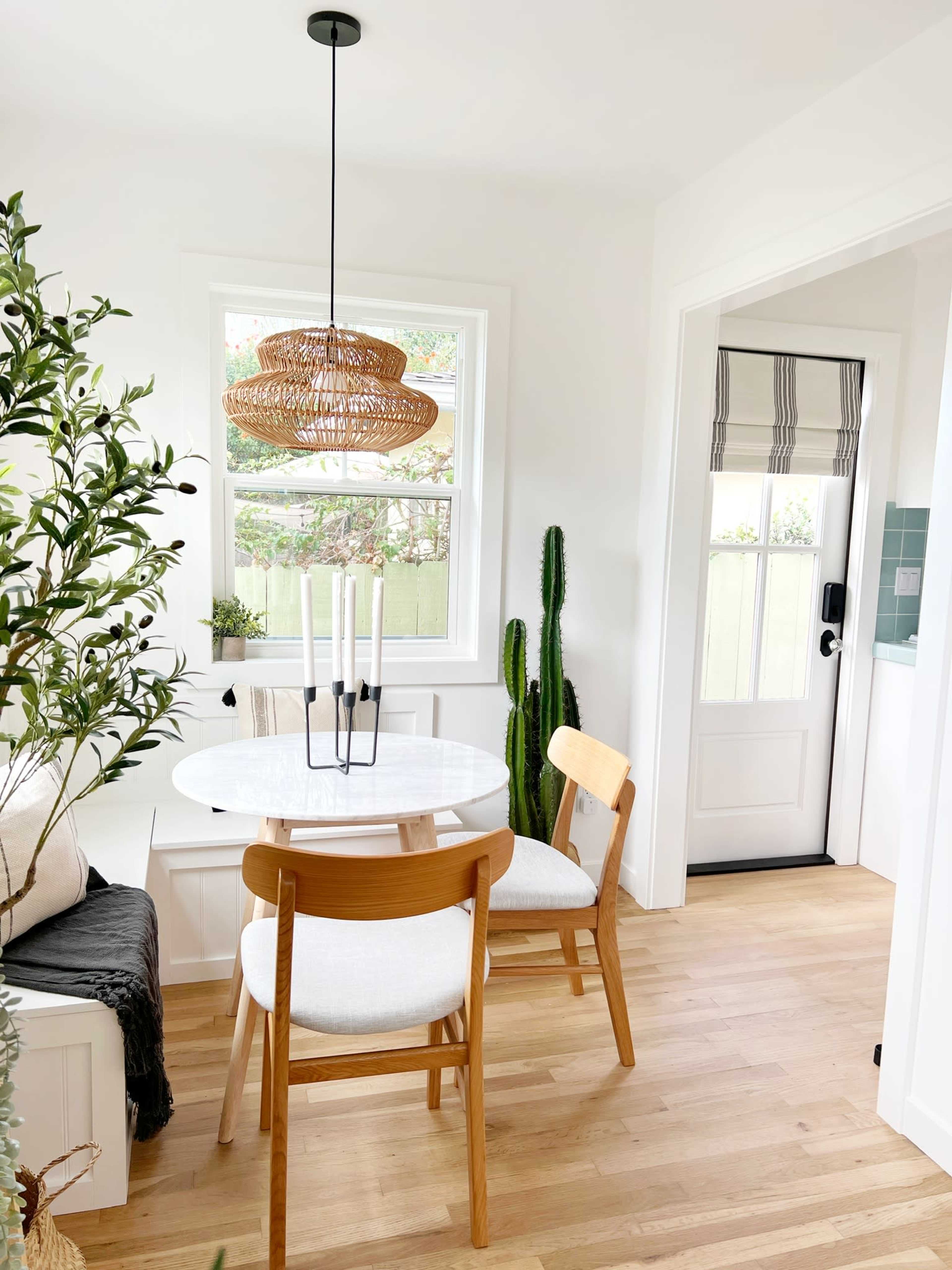 A bright dining area features a round table surrounded by wooden chairs, and a woven pendant light hangs overhead next to a window with cacti and greenery.