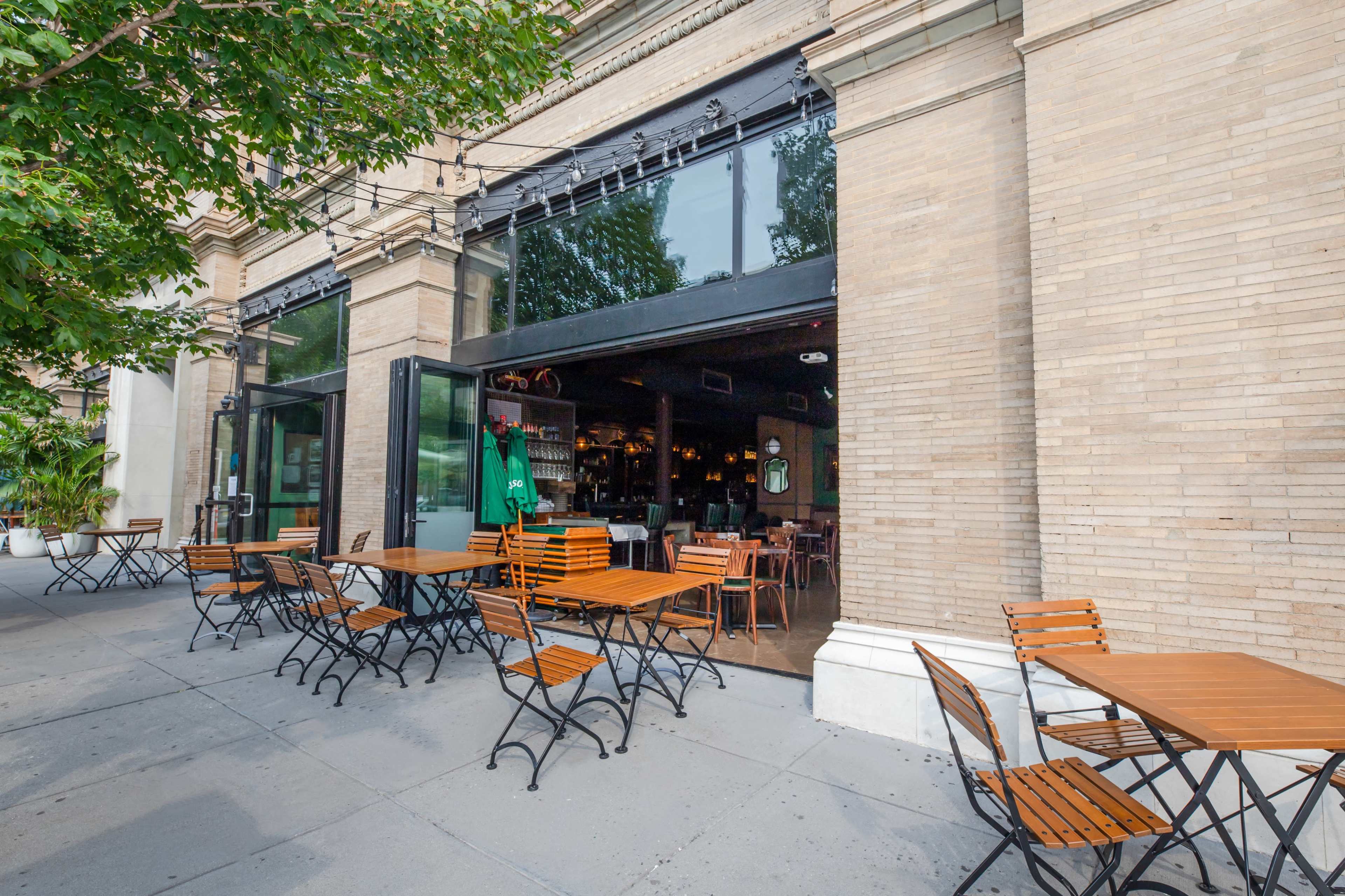 An outdoor seating area with wooden tables and chairs is situated in front of a restaurant with large open windows.