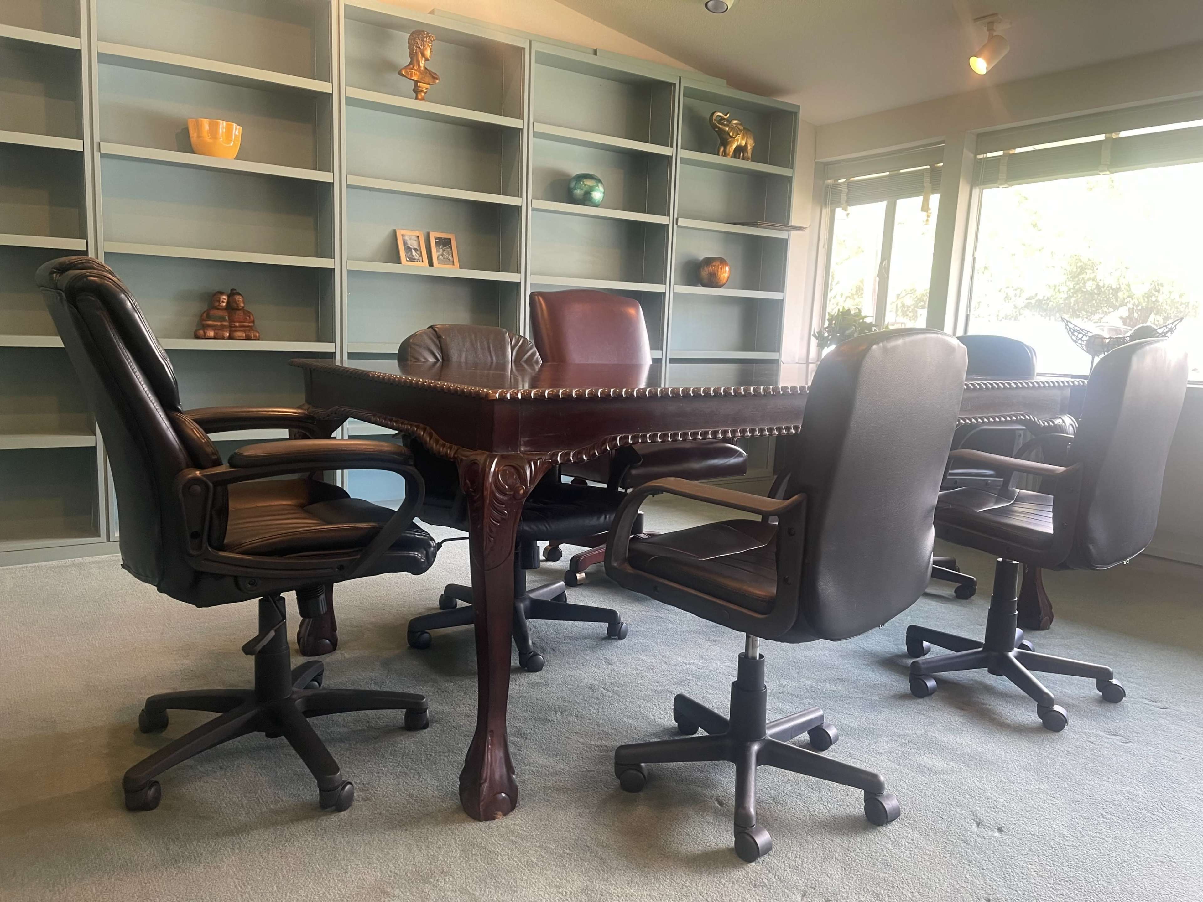 A large, ornate conference table is surrounded by five ergonomic office chairs in a well-lit room with shelves in the background.