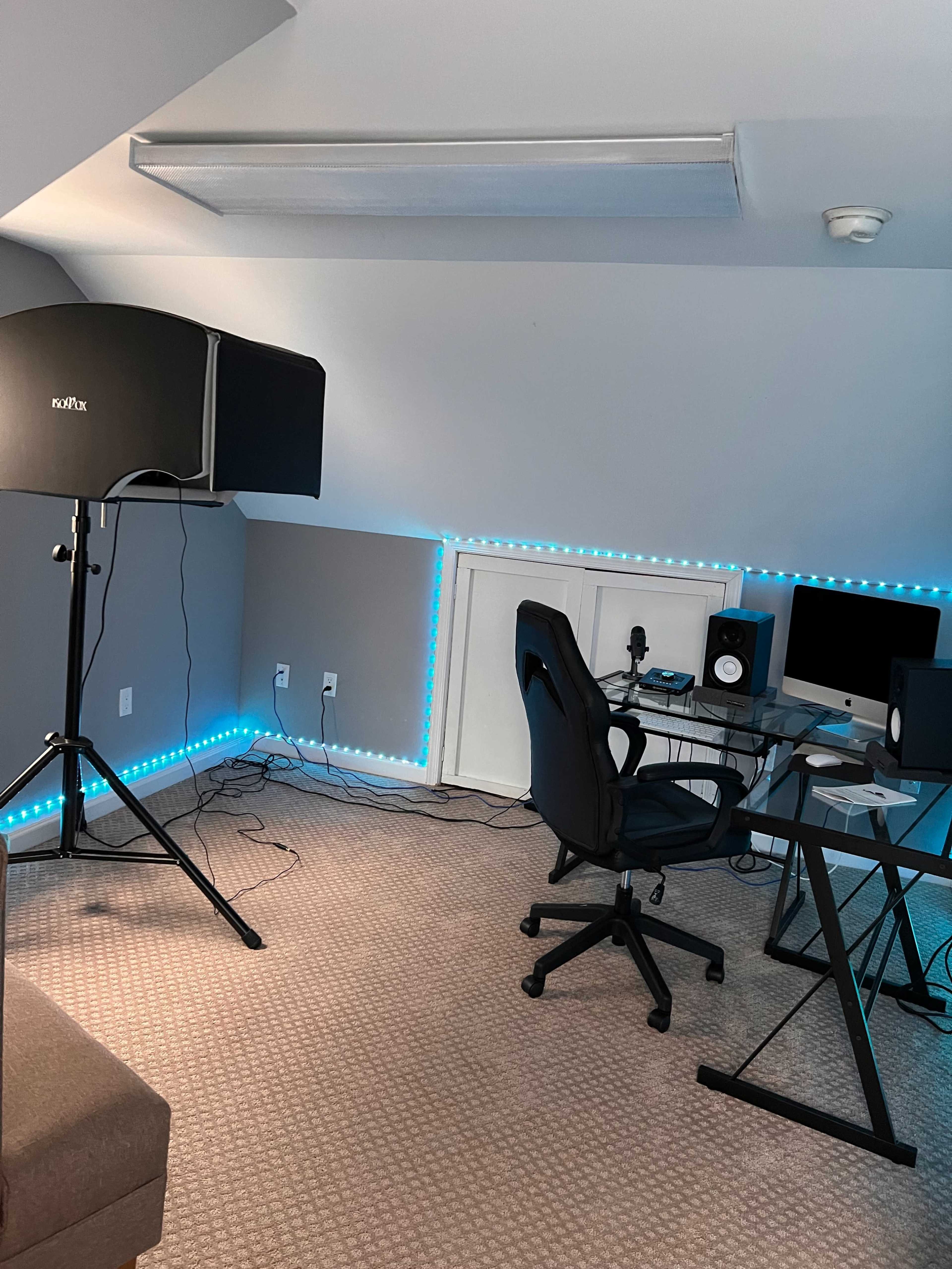 A small, modern studio space with a microphone stand, an office chair, a desk with audio equipment, and LED lighting along the walls.