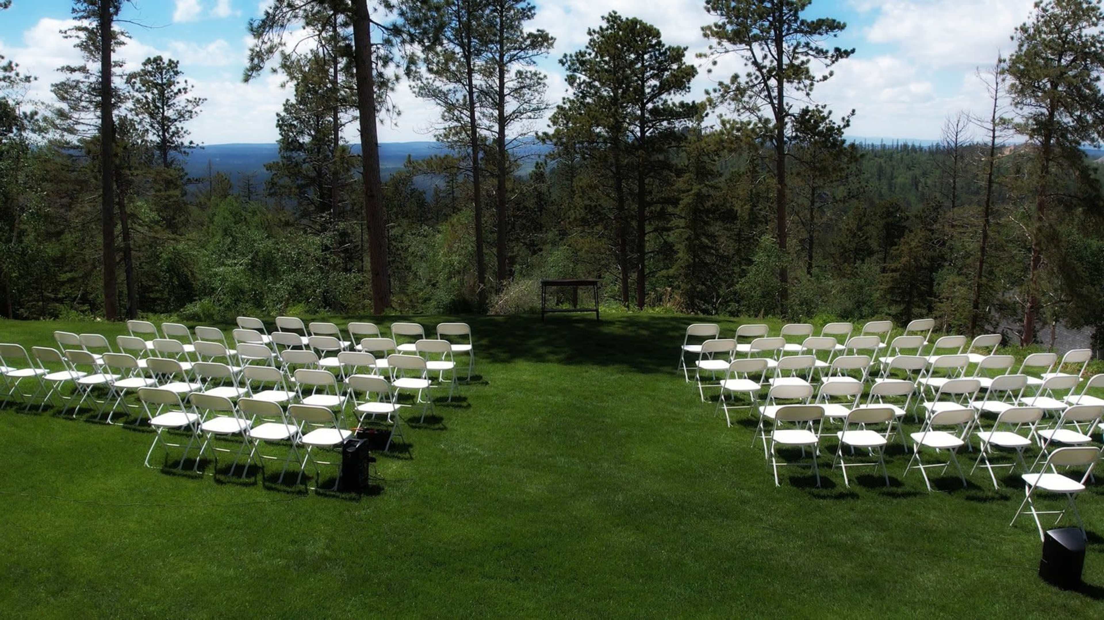 A set of white chairs is arranged in rows on a grassy area overlooking a forested landscape.