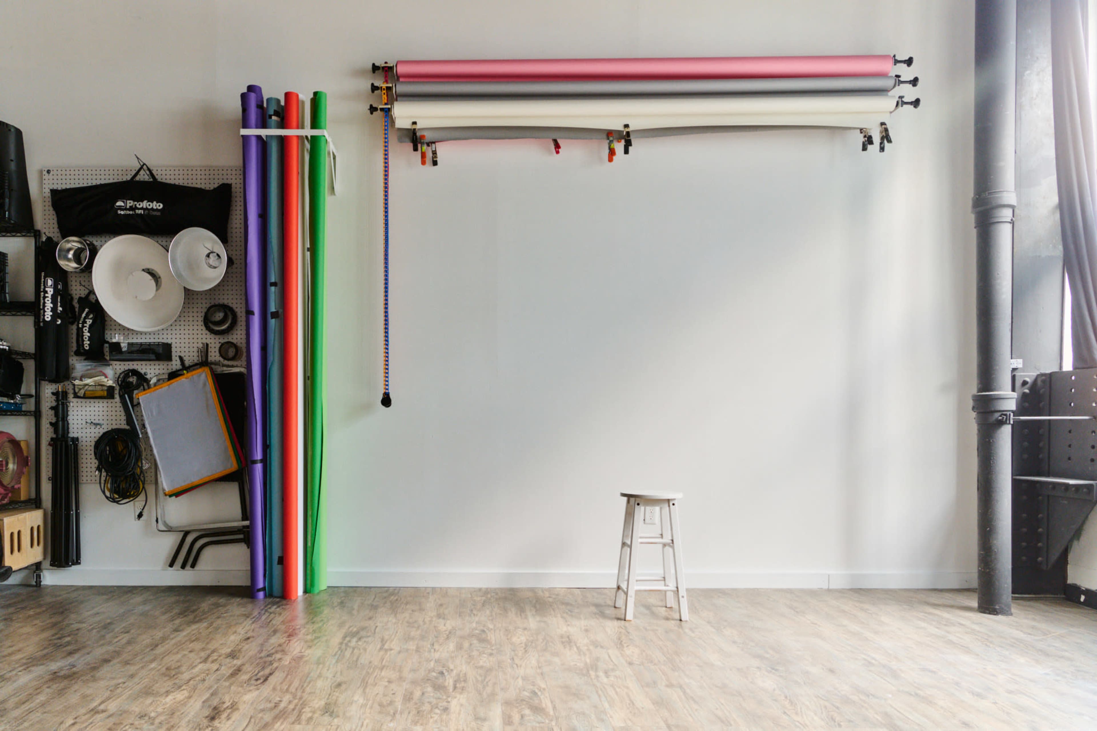 A white wall features rolls of colored backdrops hanging above a wooden floor with a single white stool positioned in the center.