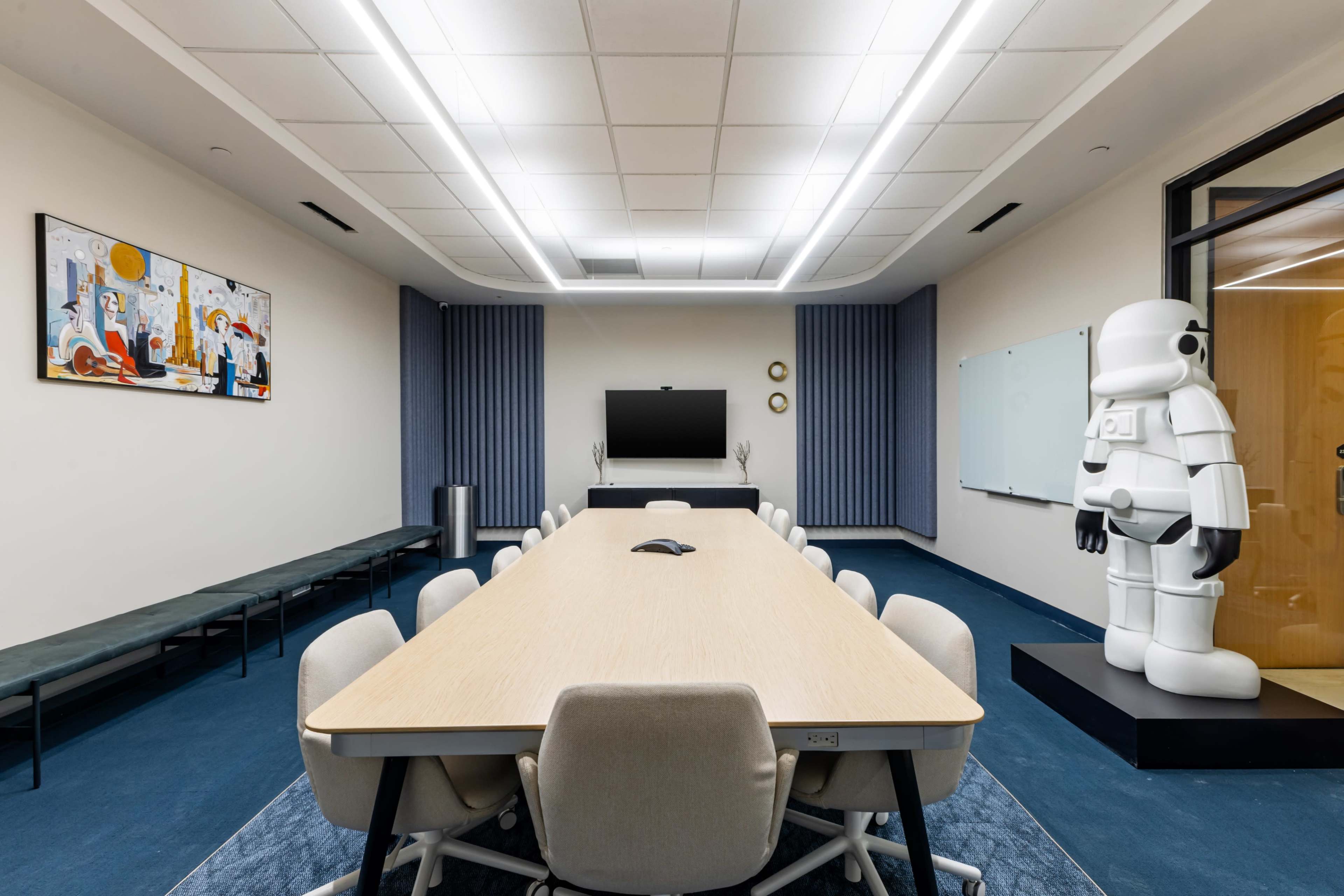 The image shows a modern conference room featuring a long wooden table, chairs, a large screen, and a life-sized astronaut figure standing in a corner.