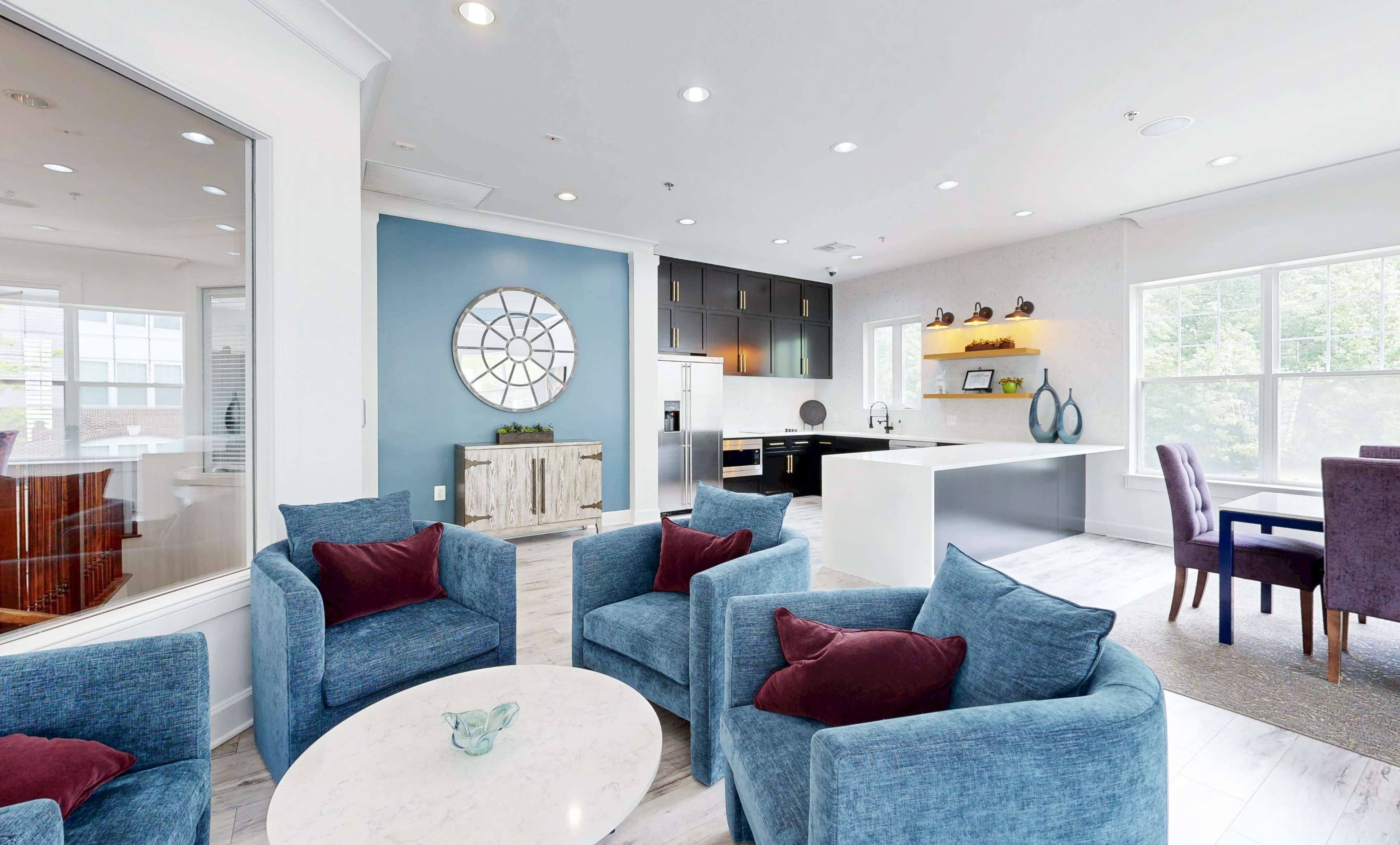 The image shows a contemporary lounge area featuring blue upholstered chairs, a circular coffee table, and a modern kitchen with white cabinetry and a dark accent wall.