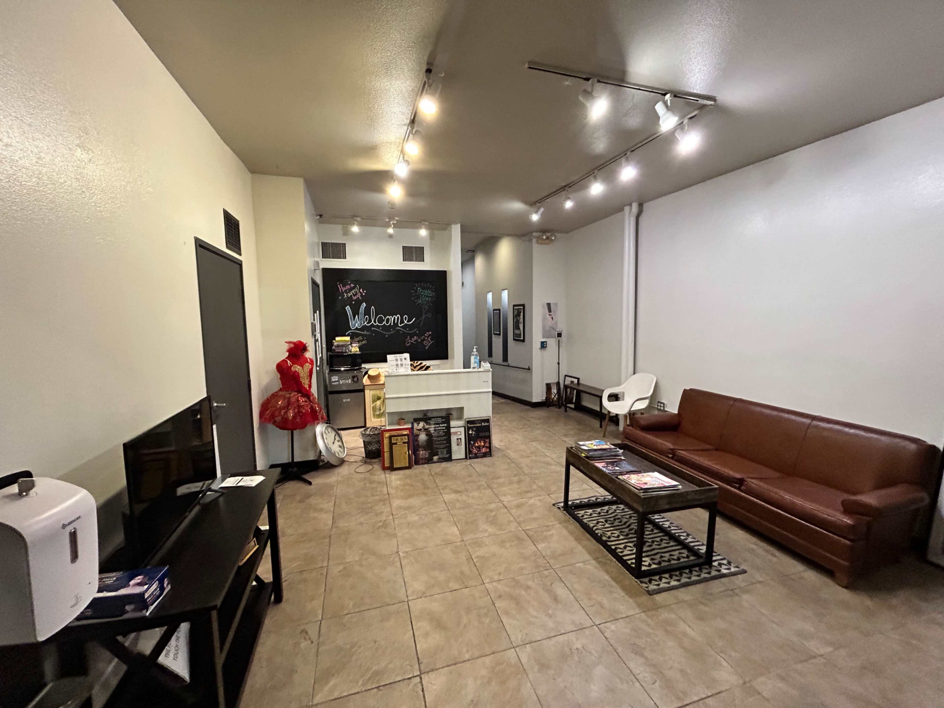 The image shows a clean lobby area with a reception desk, a brown sofa, a coffee table, and a mannequin dressed in a red outfit.