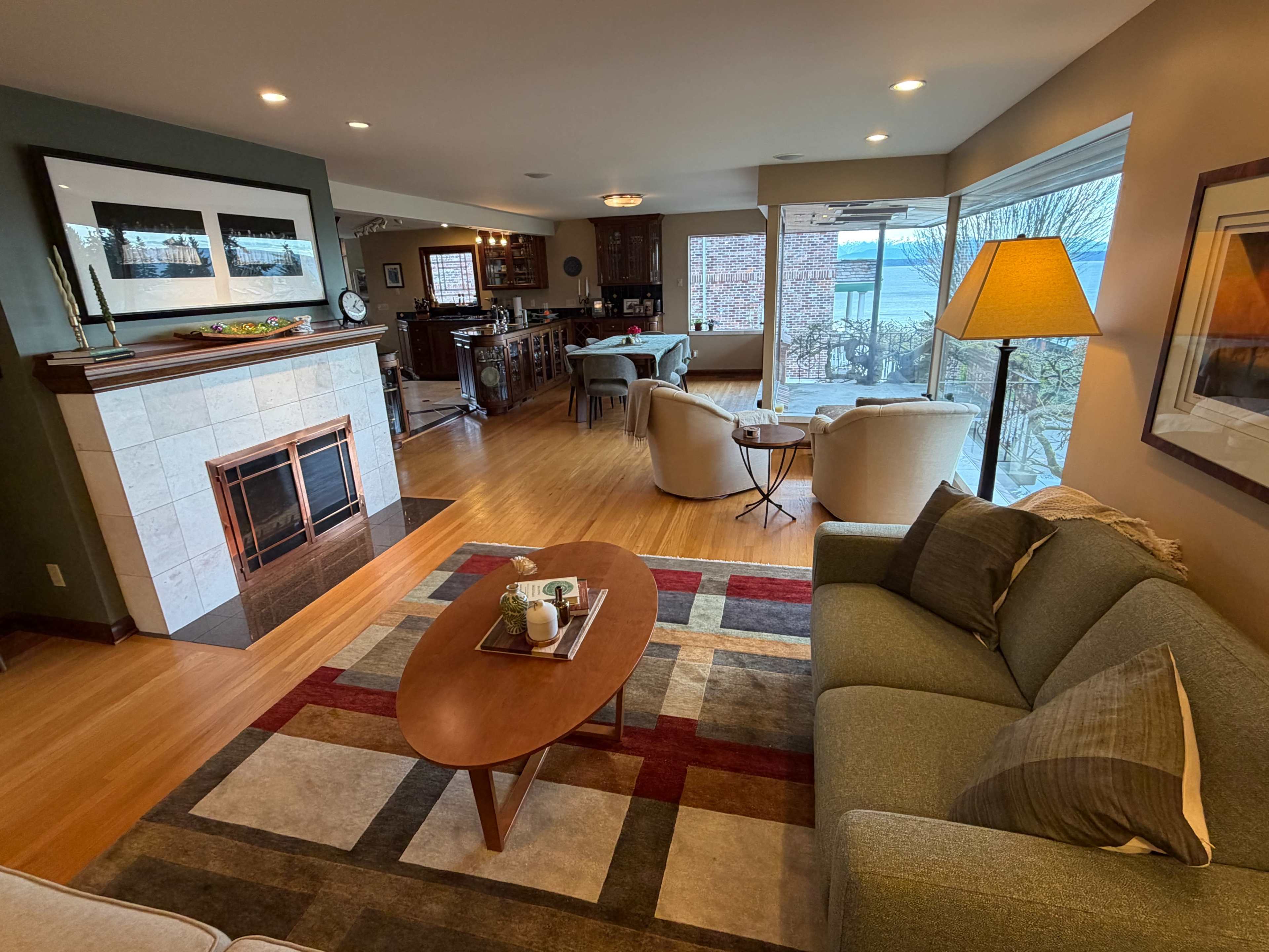 The image depicts a cozy living room featuring a sofa, a coffee table, and a stone fireplace, with large windows overlooking an outdoor patio.