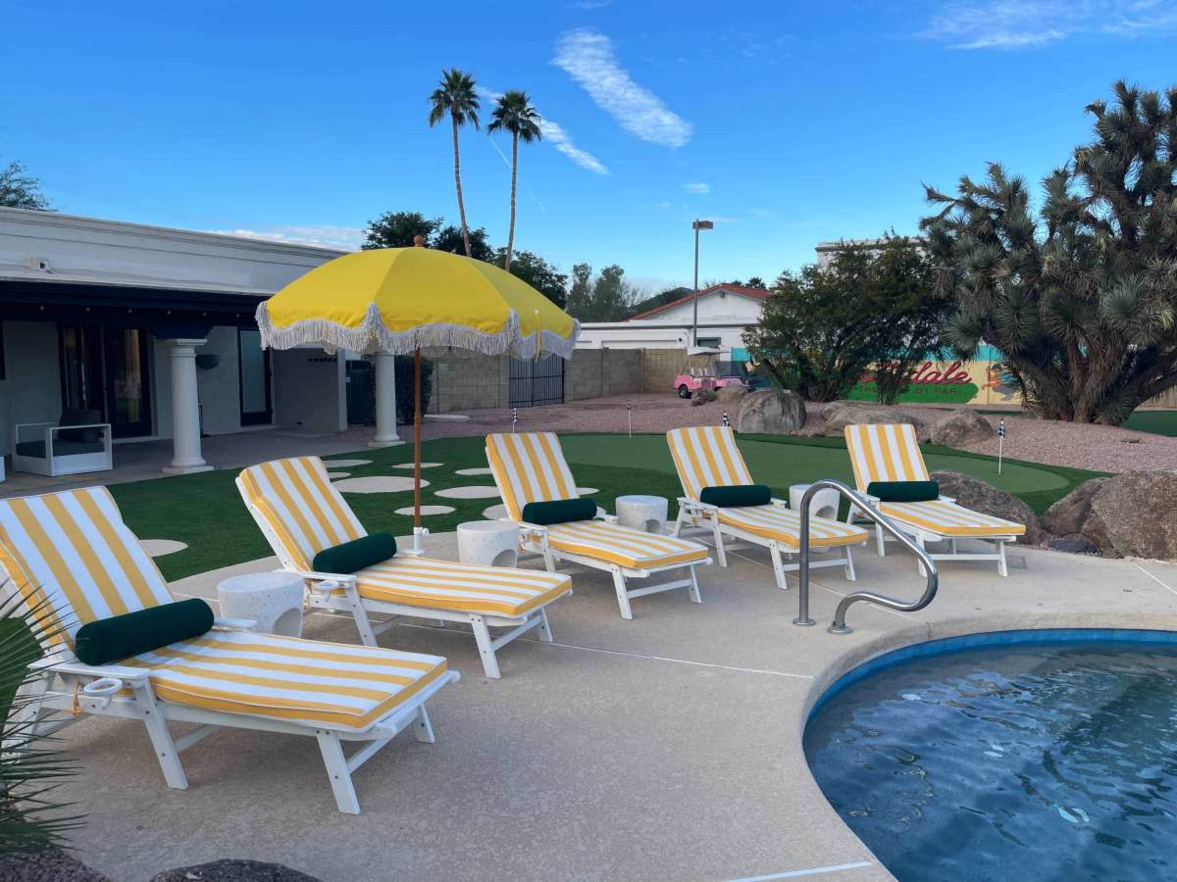 The image shows a poolside area with four yellow and white striped lounge chairs, a yellow umbrella, and a pool surrounded by landscaping.