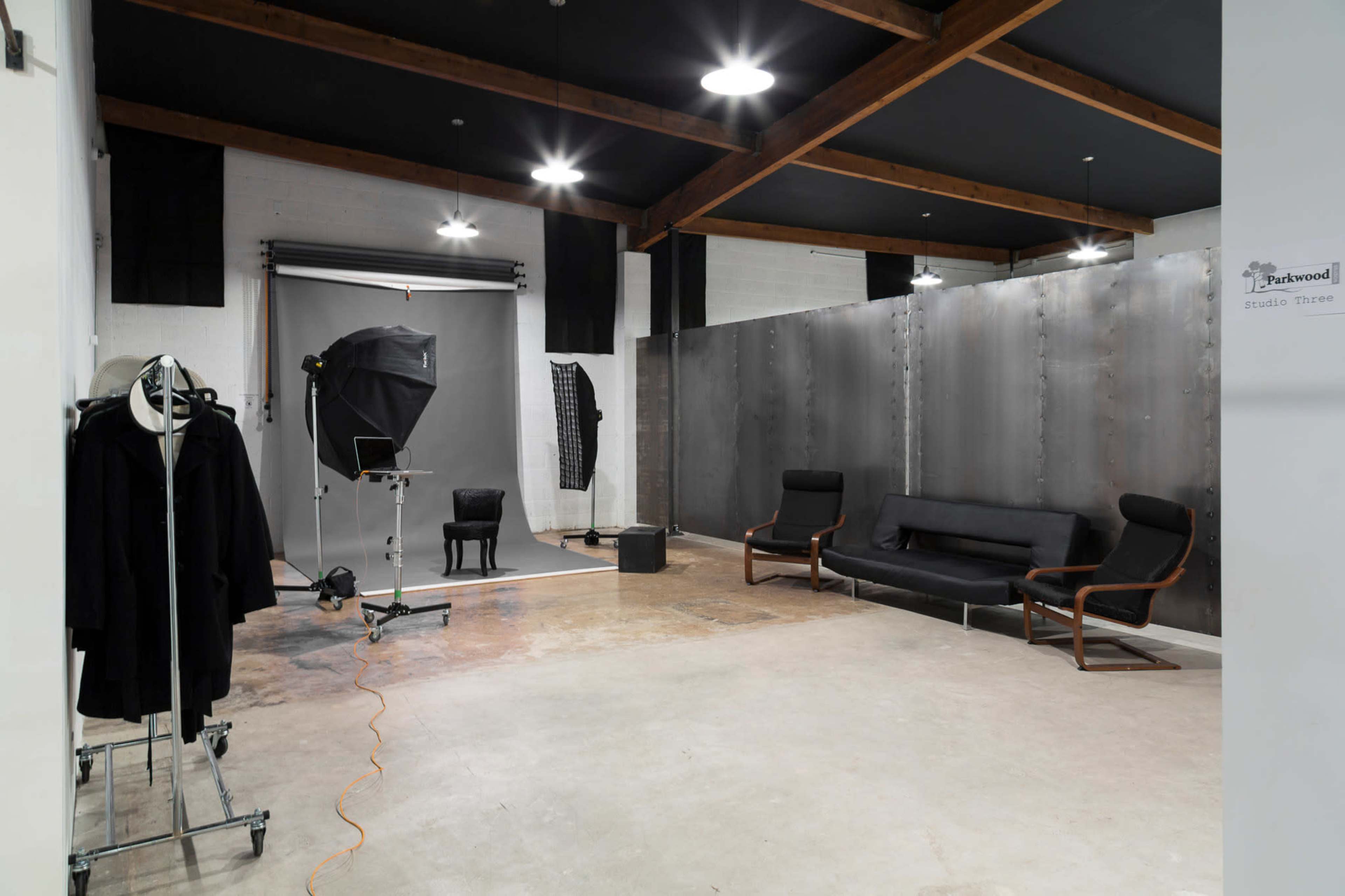 A spacious photography studio featuring a gray backdrop, a lighting setup, a black chair, and a sofa area with minimalist decor.
