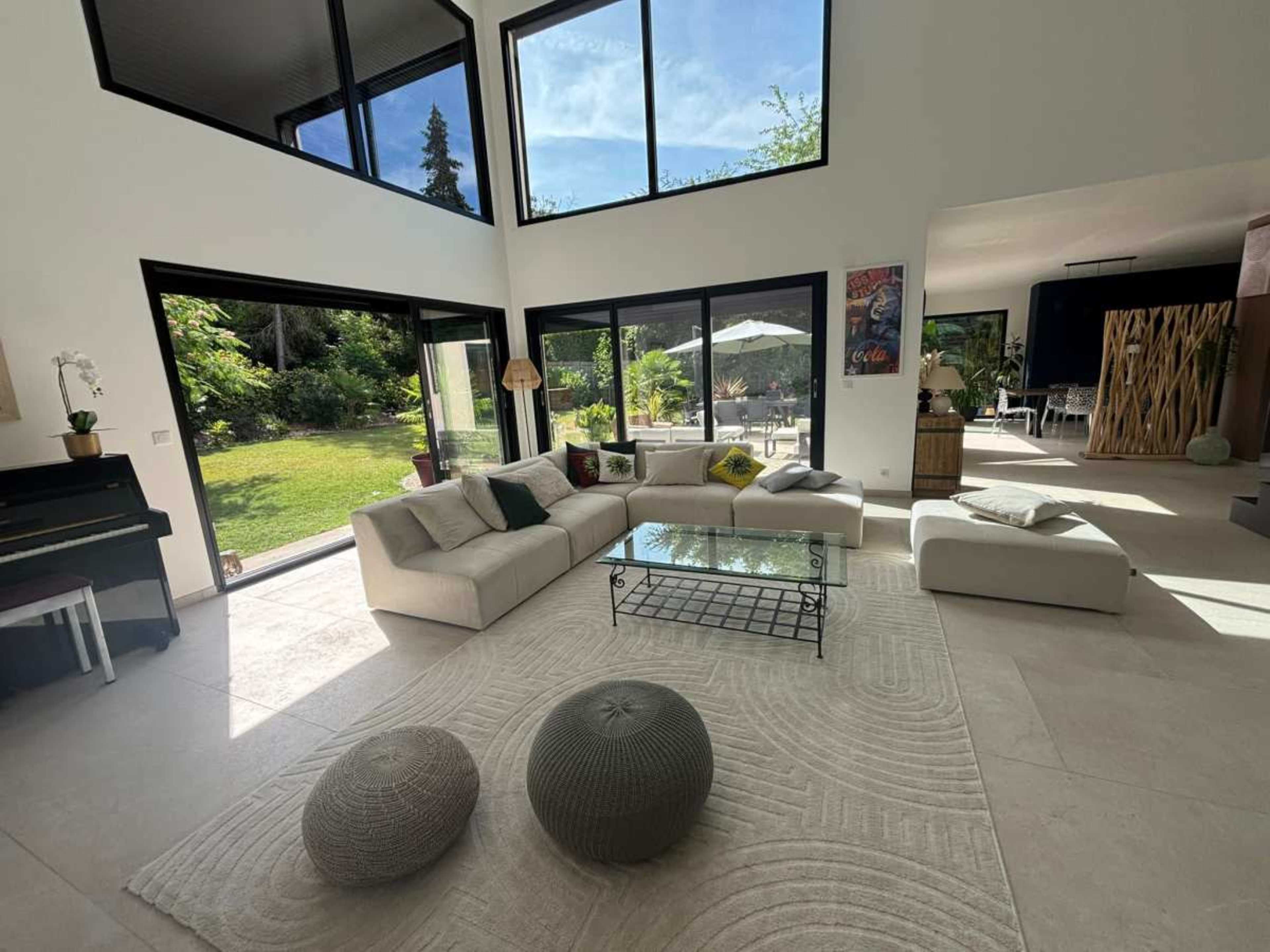 The image shows a spacious living room with a large sectional sofa, a glass coffee table, and large windows overlooking a garden.