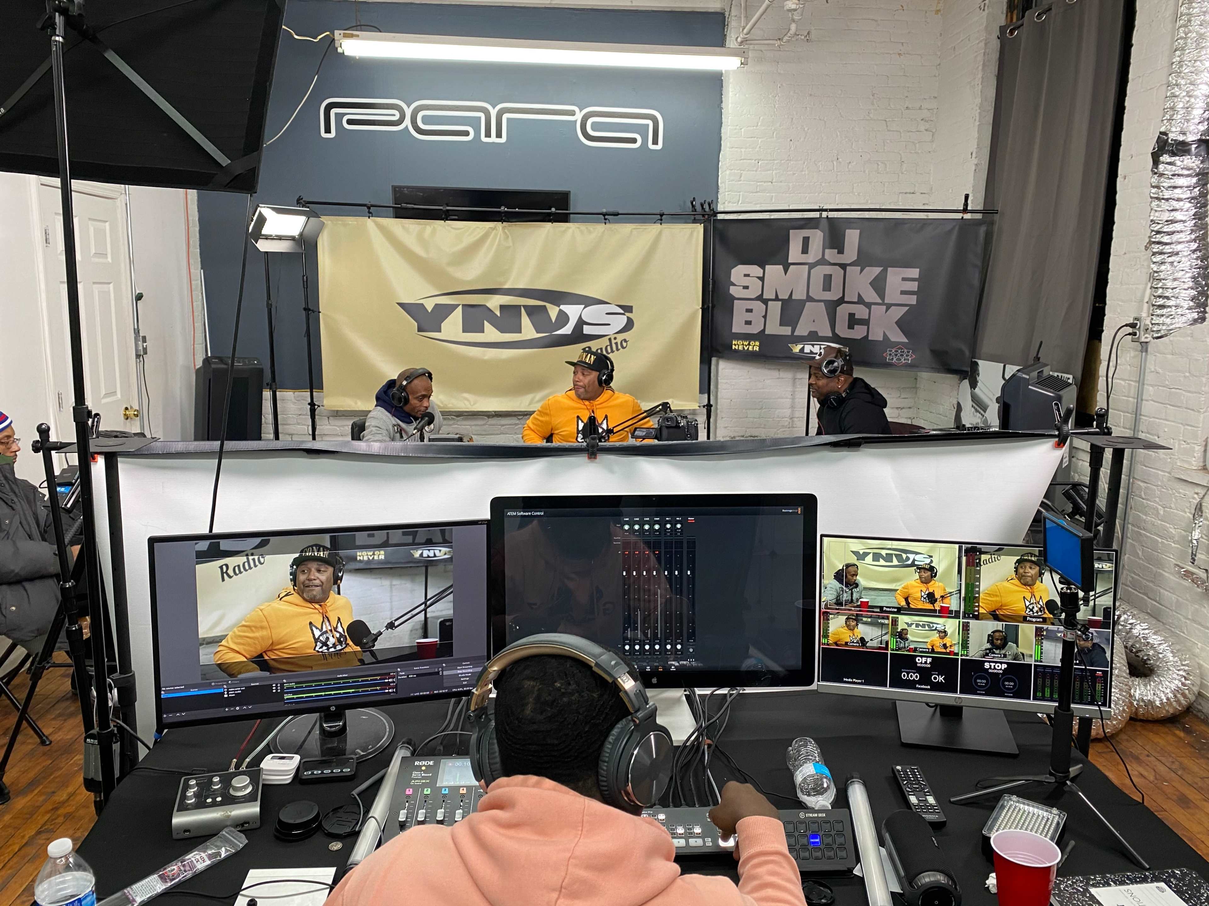 A radio studio setup with three individuals engaged in conversation, surrounded by audio equipment and banners displaying the station's name and DJ's tag.