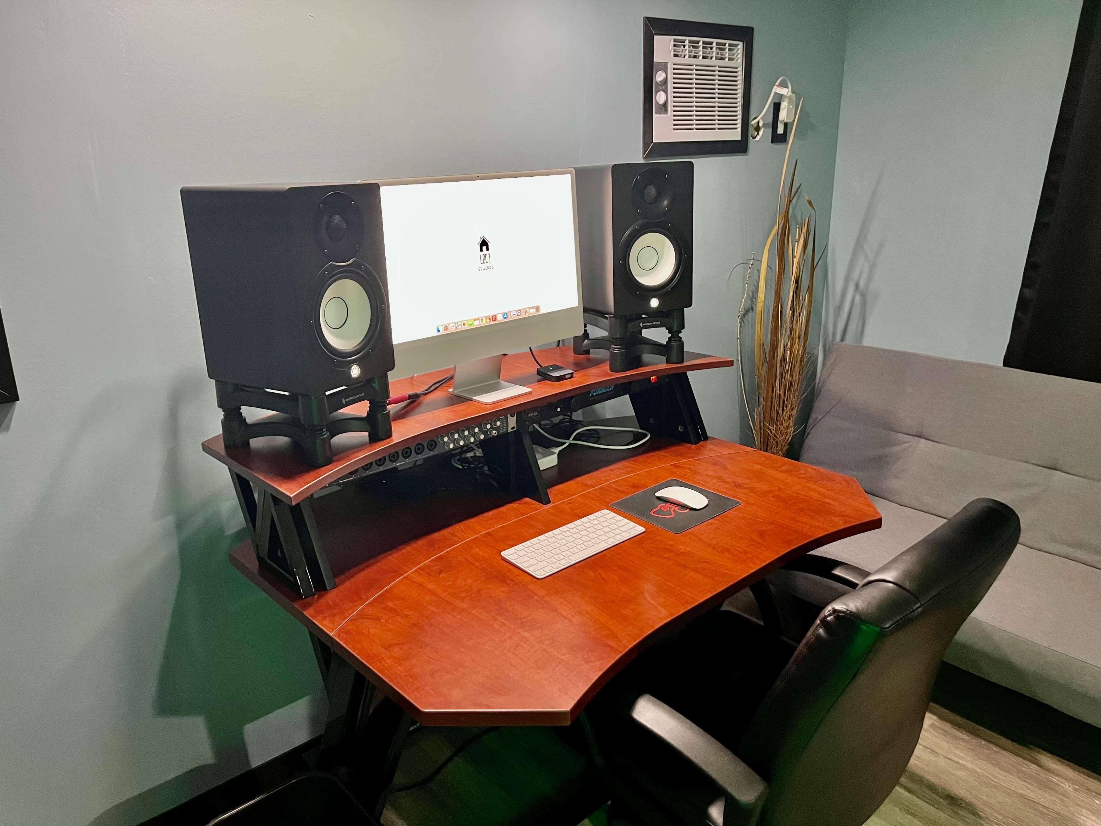 A modern workspace with a computer monitor, studio speakers on elevated stands, a keyboard, and a comfortable chair in a room featuring green walls and a gray couch.