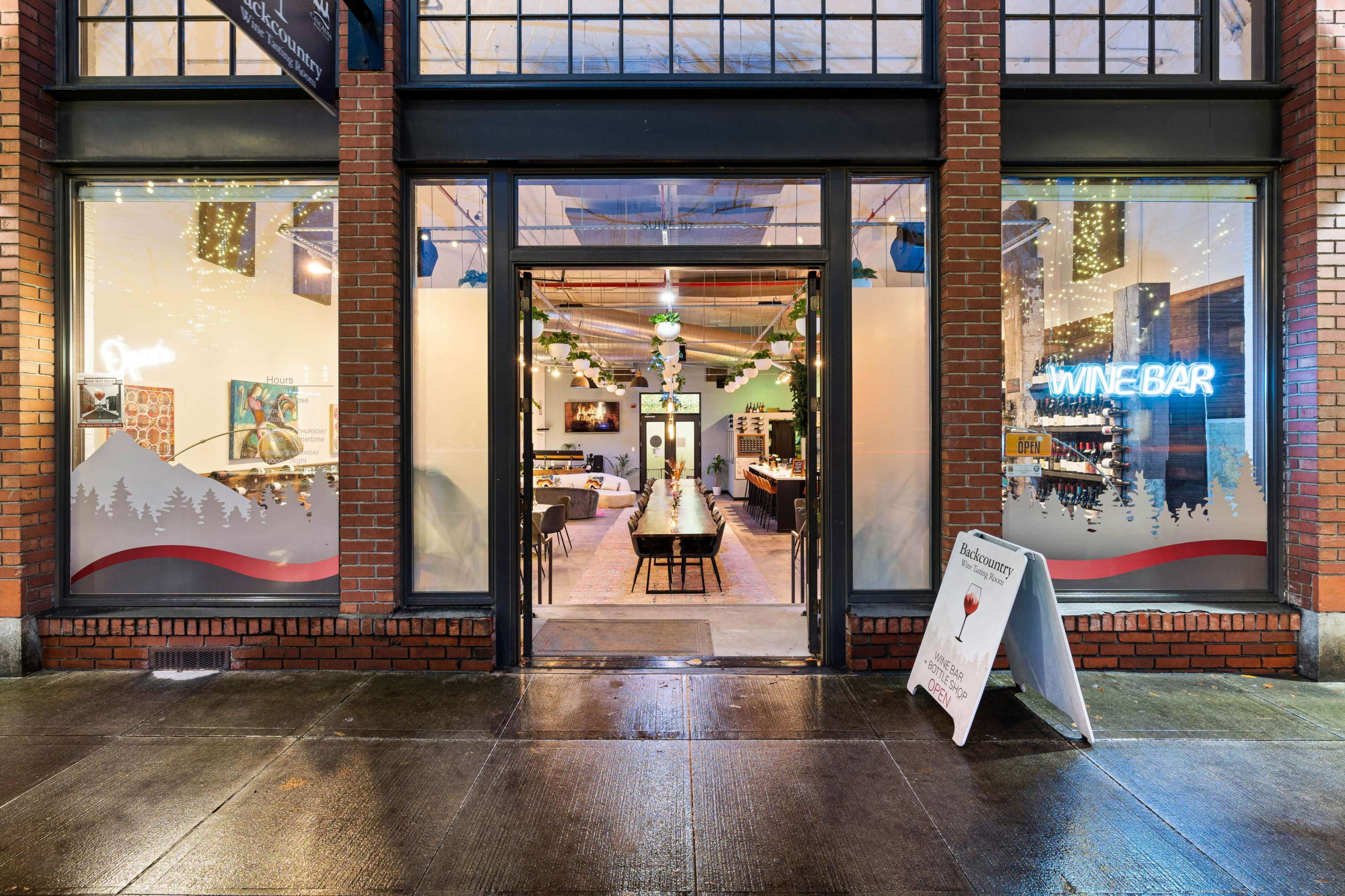 A wine bar with large front windows displays a modern interior, featuring wooden tables and decorative lights, under a brick facade.