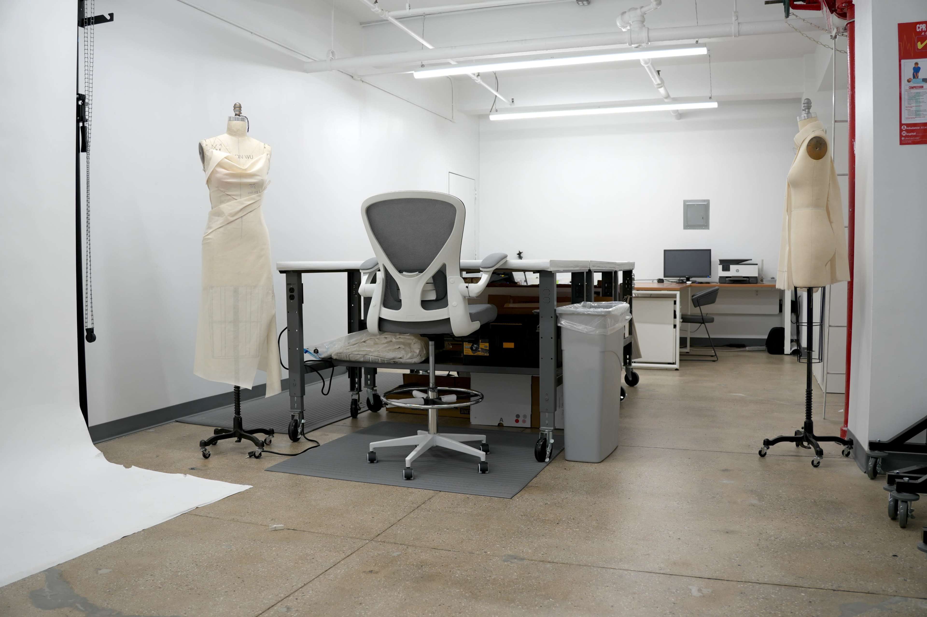 The image shows a minimalist work studio with a central desk, an ergonomic chair, and two dressmaker forms on stands.