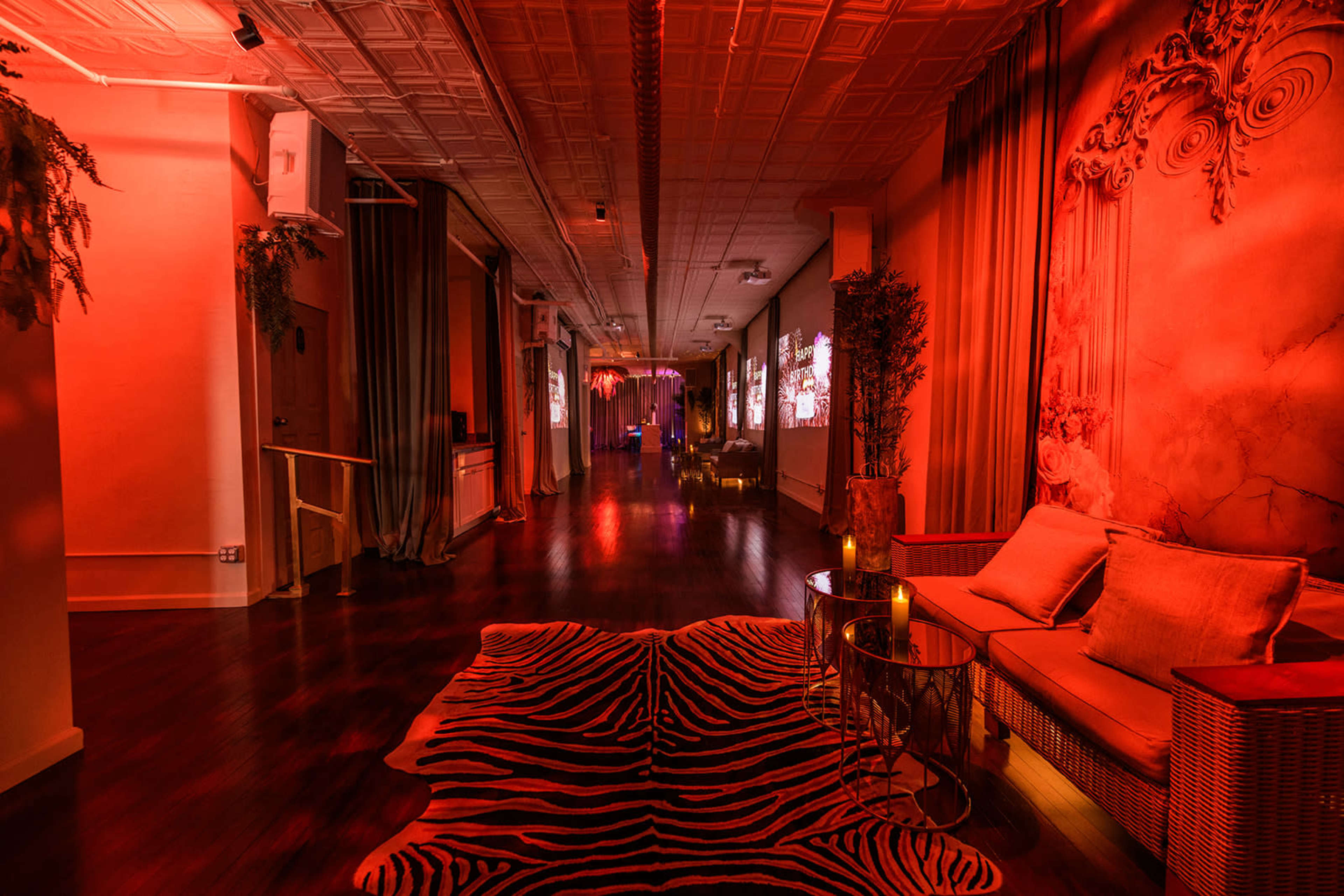 The image shows a long hallway with red lighting, featuring a zebra-striped rug, a seating area with wicker furniture, and decorative elements including potted plants.