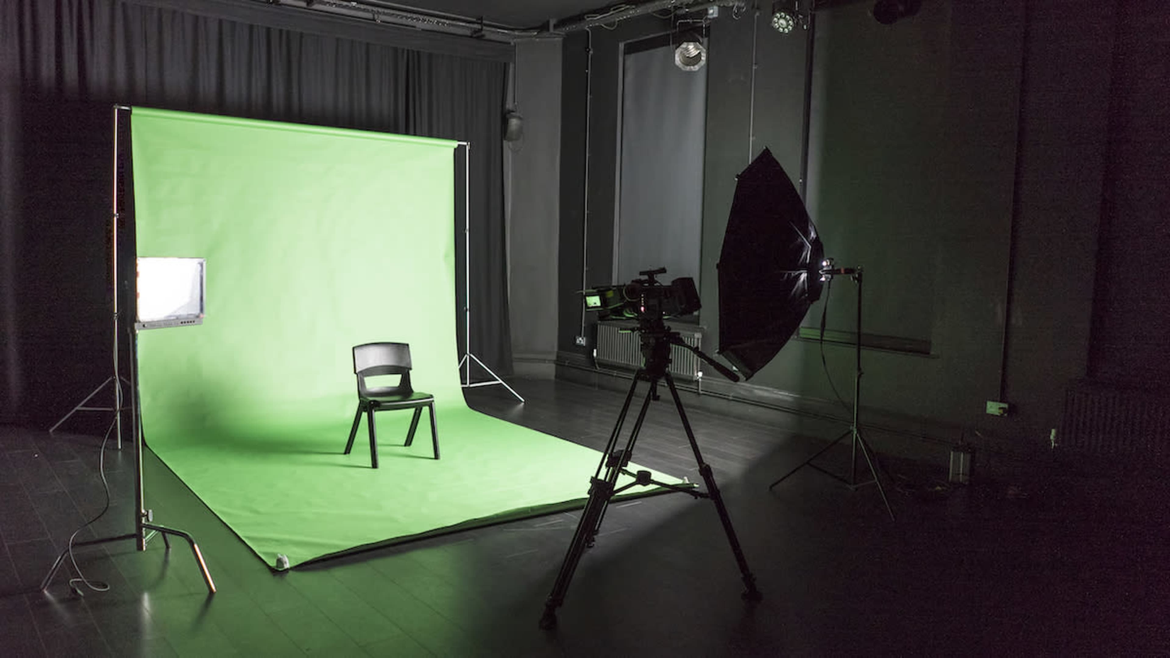 The image shows a photography studio set up with a green backdrop, a chair in the center, and a video camera on a tripod pointed towards the chair.