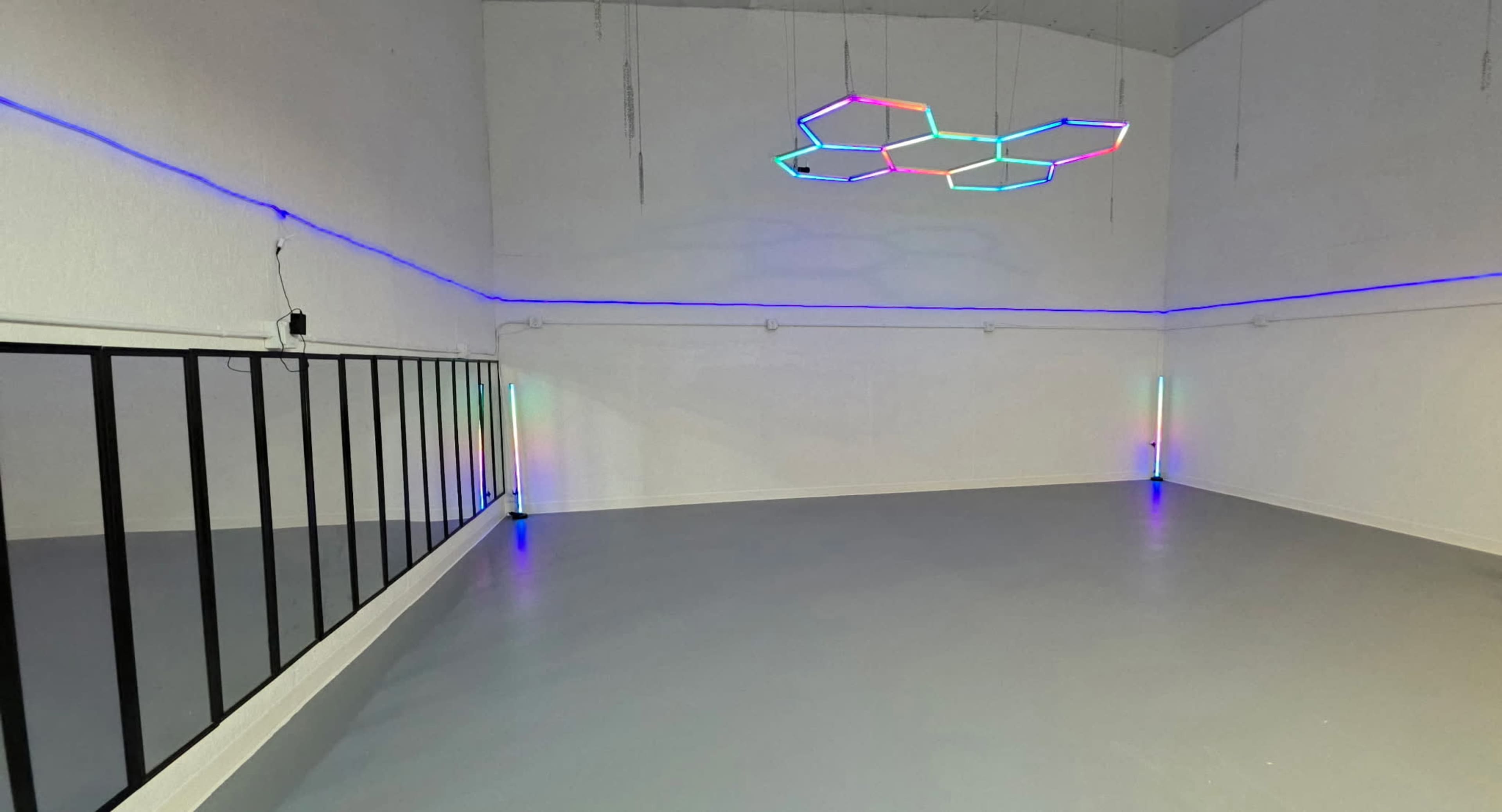 The image shows a minimalist room with gray flooring, illuminated by colorful LED lights along the walls and a geometric light fixture hanging from the ceiling.