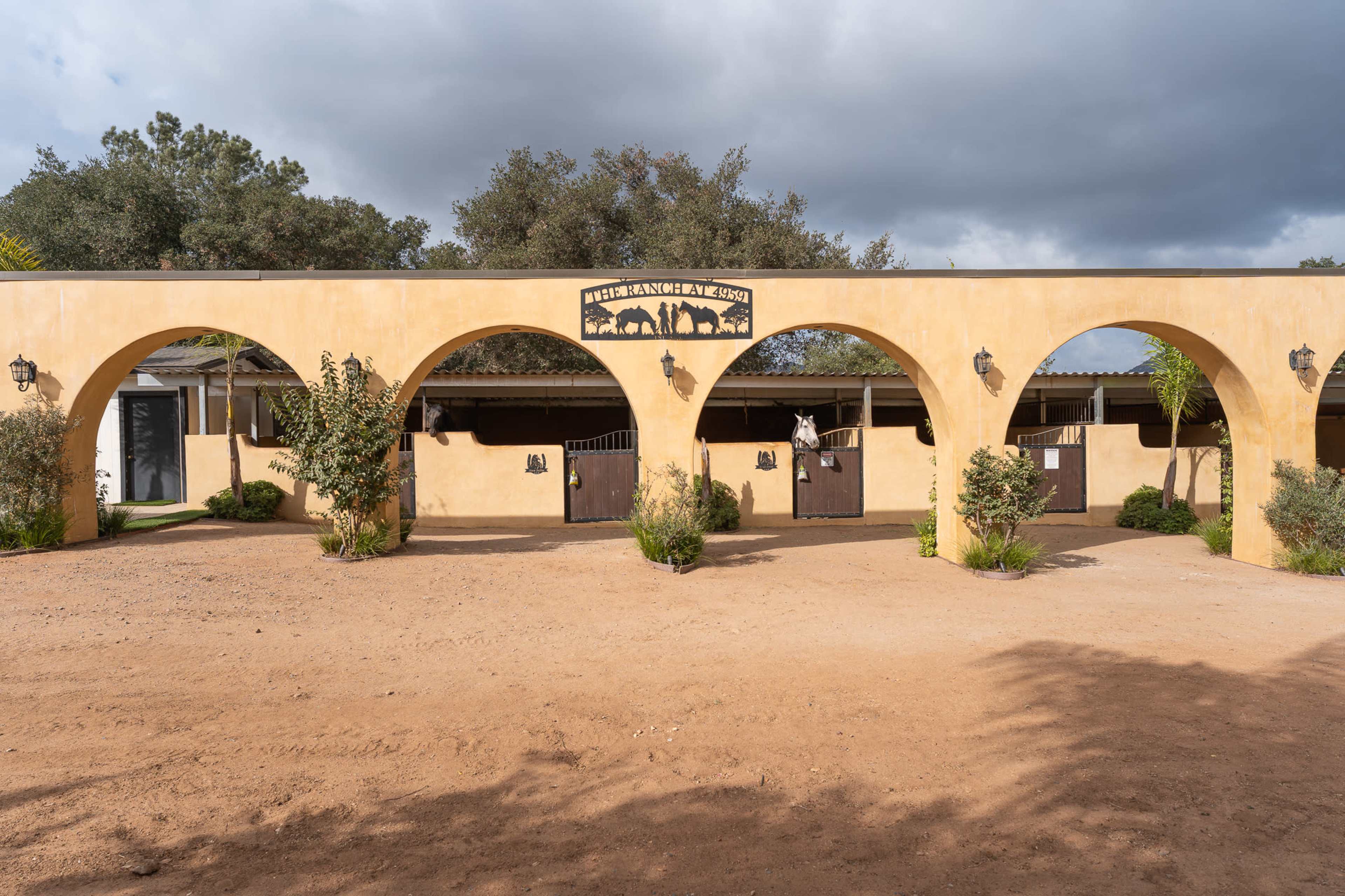 The image depicts a stable structure with archways, featuring several horse stalls and landscaping on a dirt pathway under a cloudy sky.