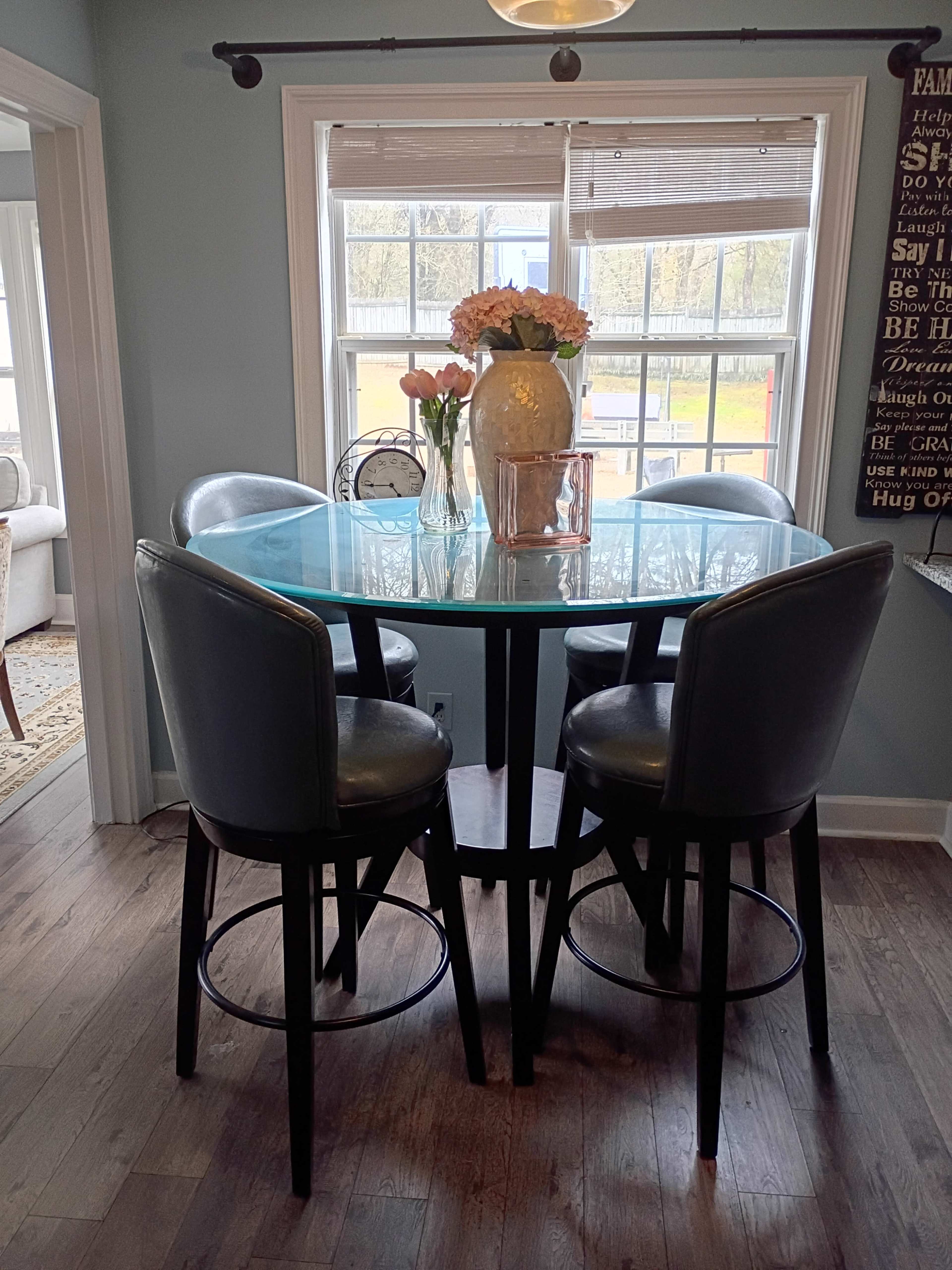 A round glass-top table with four black leather bar stools is set near a window, adorned with a vase of flowers and a decorative bowl.