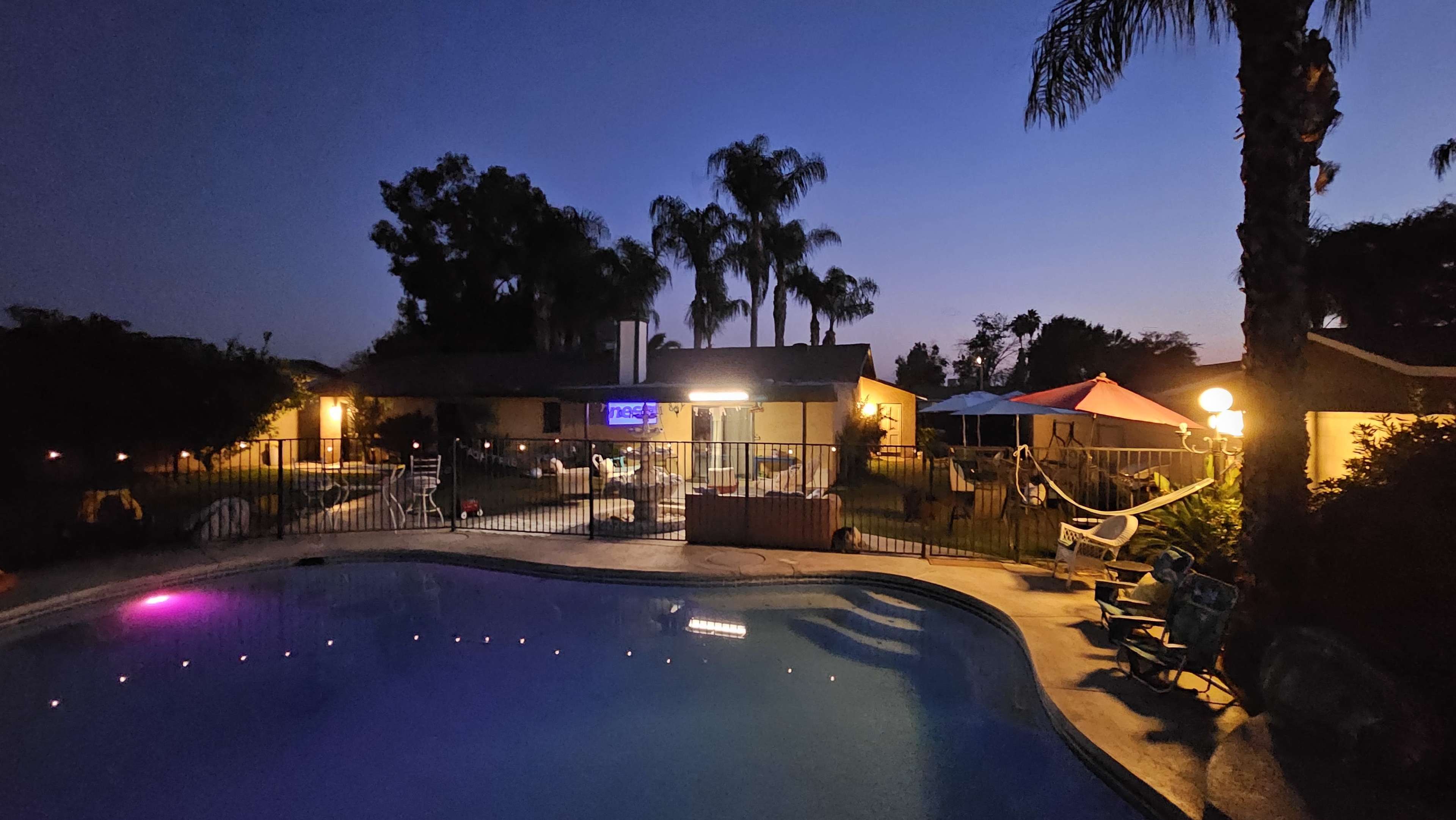 The scene features a backyard pool area at dusk, surrounded by palm trees and patio furniture, with outdoor lighting illuminating the space.