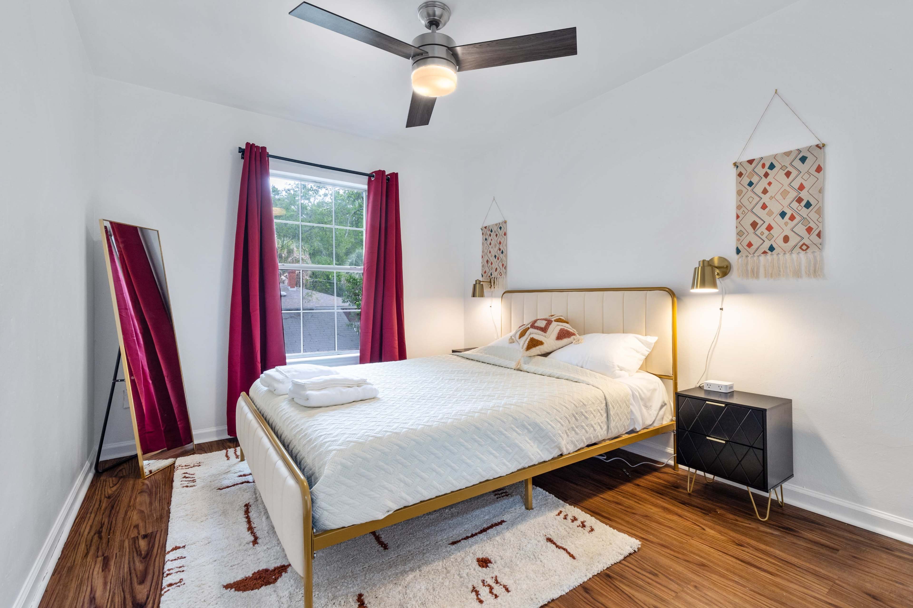A modern bedroom featuring a queen-sized bed with a white quilt, a large window with red curtains, a full-length mirror, and a black nightstand with lamps on either side.