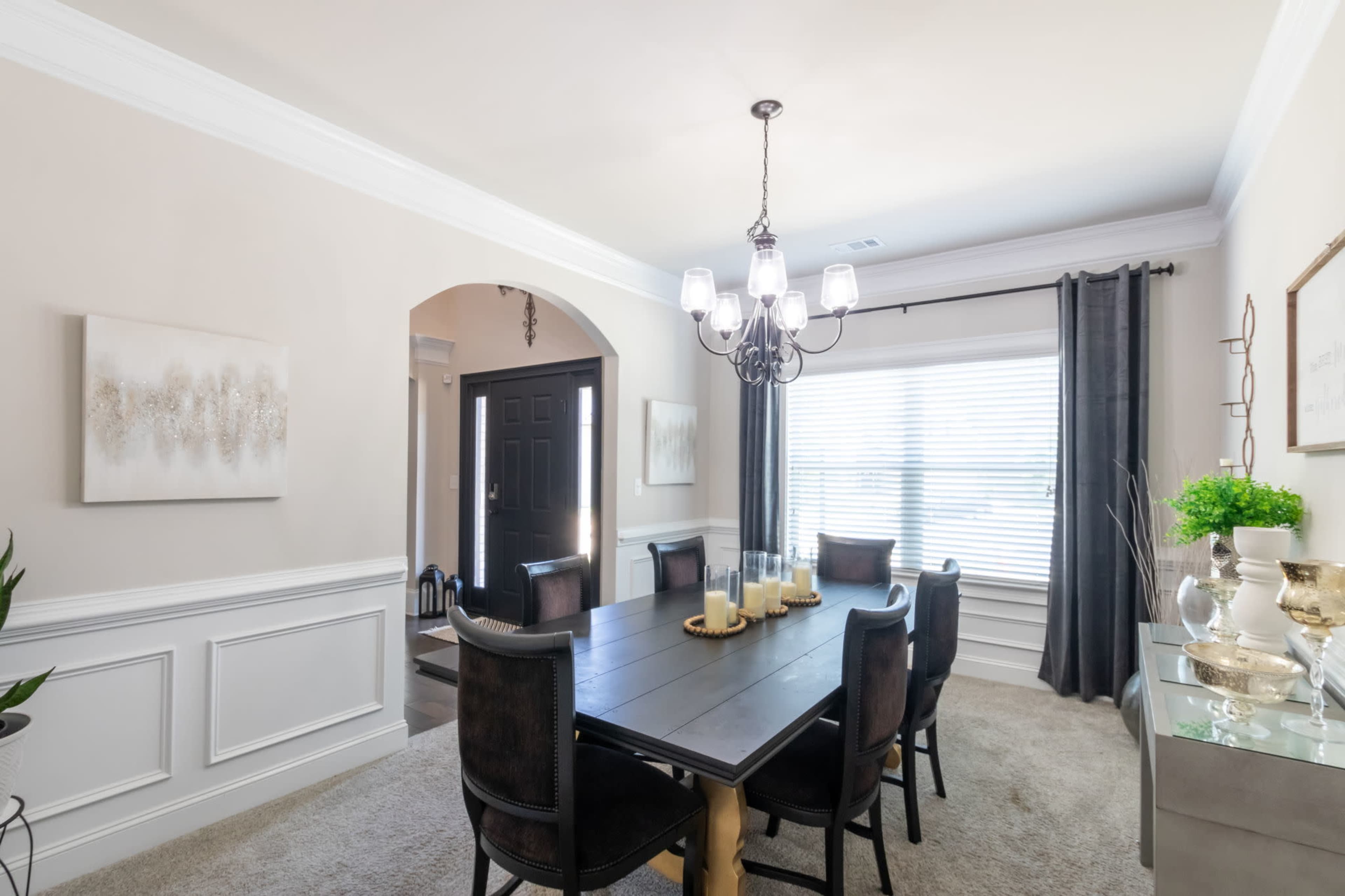 A dining room features a large table surrounded by chairs, with a chandelier overhead and large windows covered by curtains.