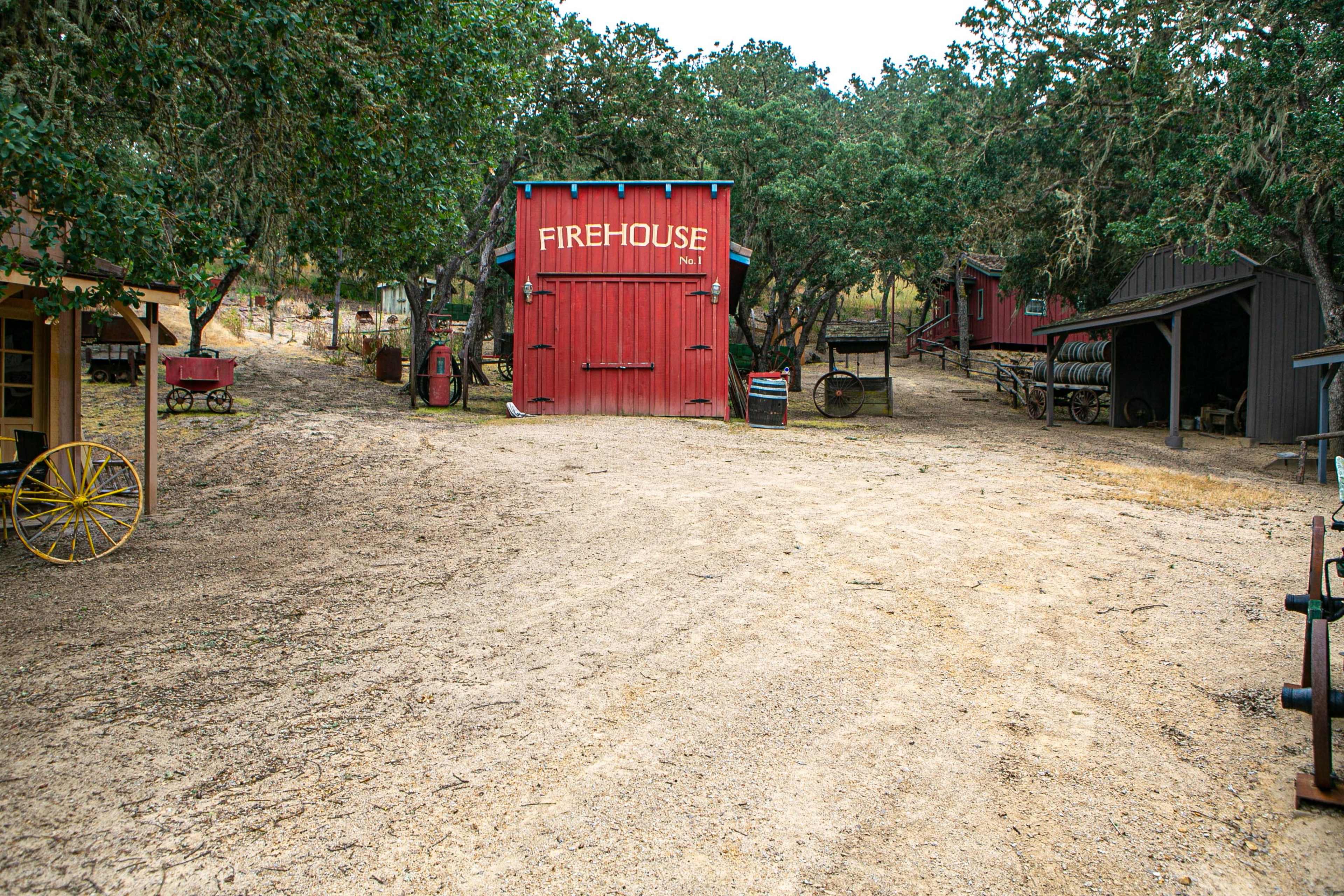 A dirt road leading to a red firehouse surrounded by wooden structures and trees.