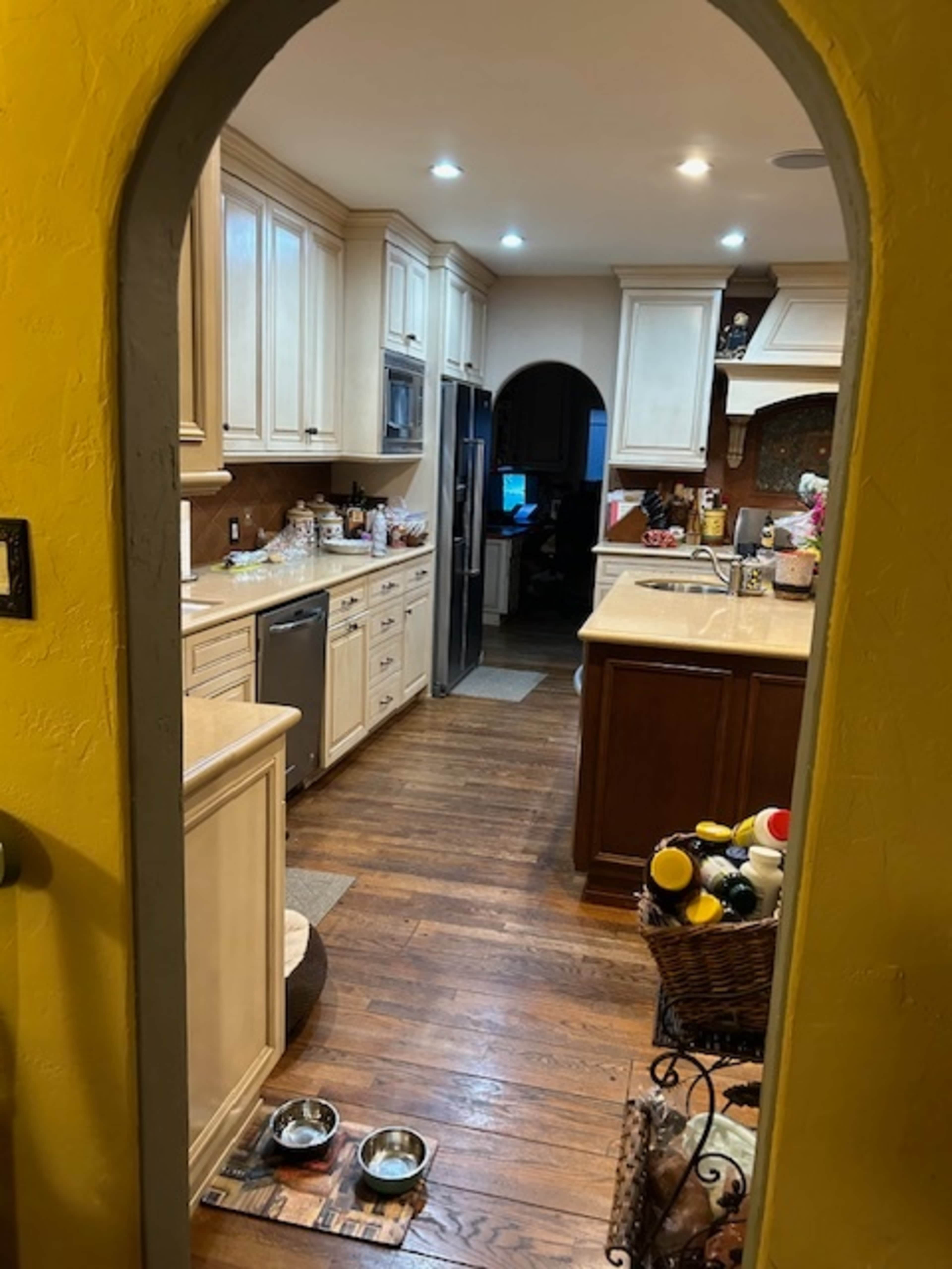 A spacious kitchen with wooden floors, light-colored cabinetry, and a view through an archway leading to another area of the home.