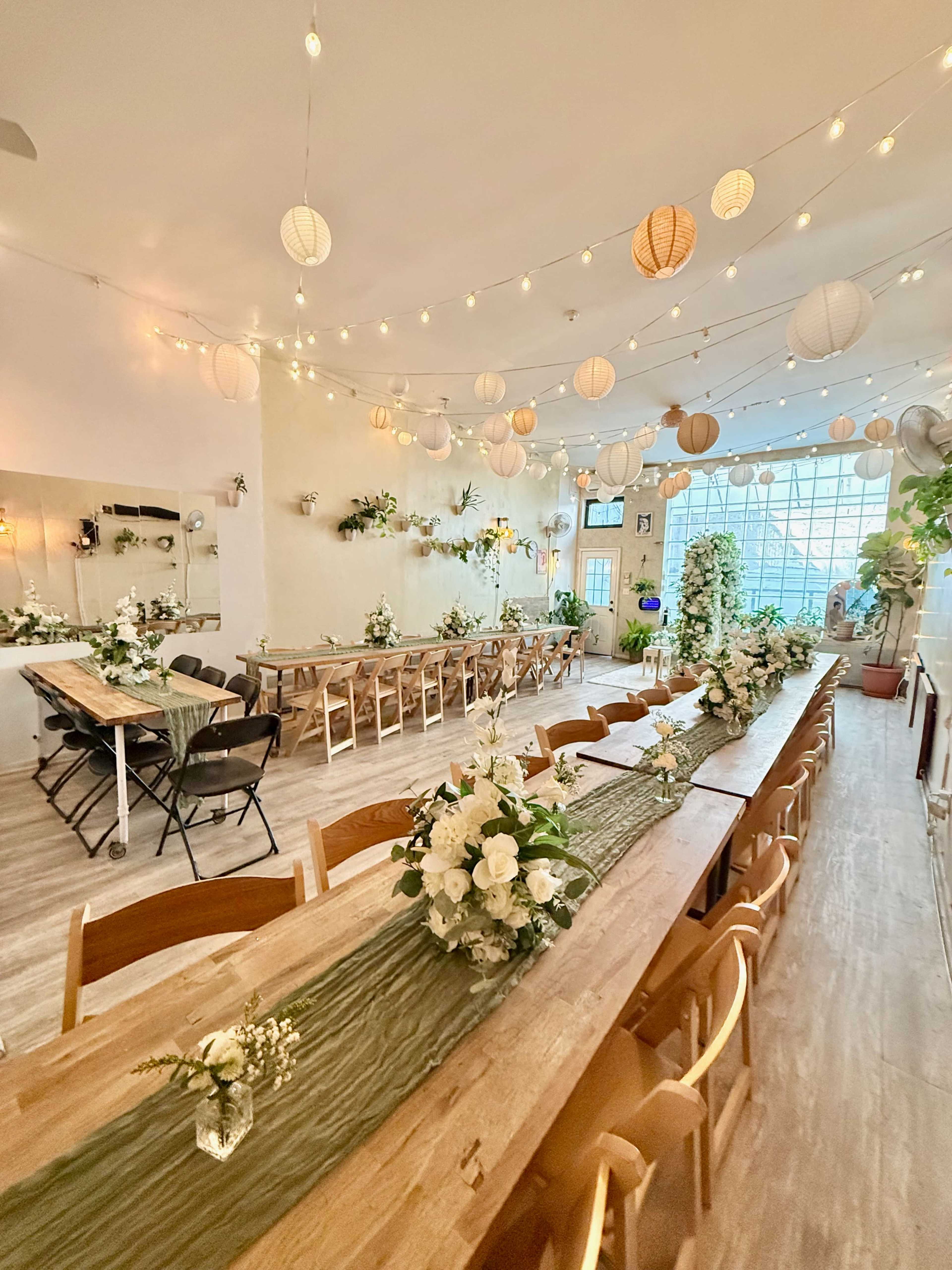 The image shows a bright, decorated indoor event space with long wooden tables arranged for dining, adorned with floral centerpieces and hanging paper lanterns.