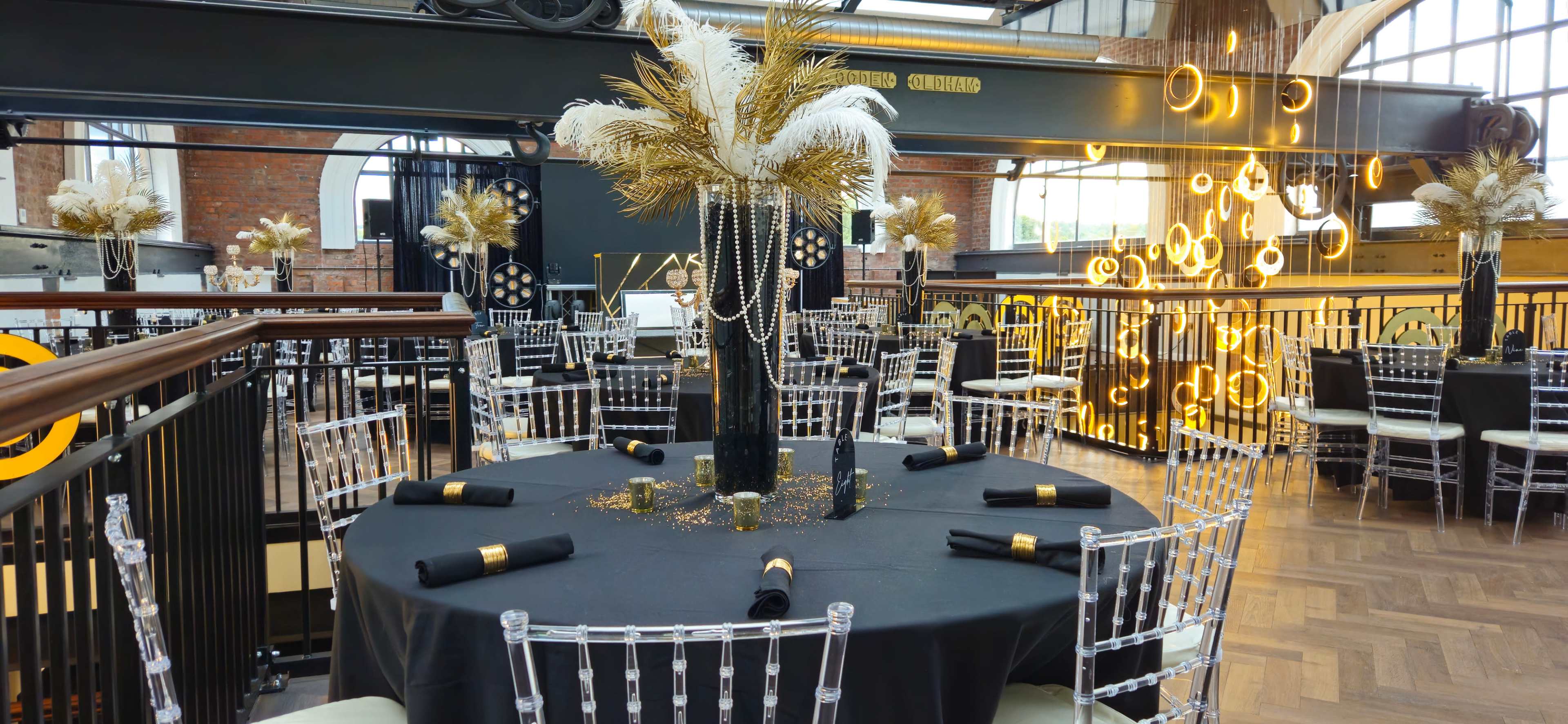 The scene shows a decorative dining area with black tables, clear acrylic chairs, and gold-themed centerpieces, all set for an event.