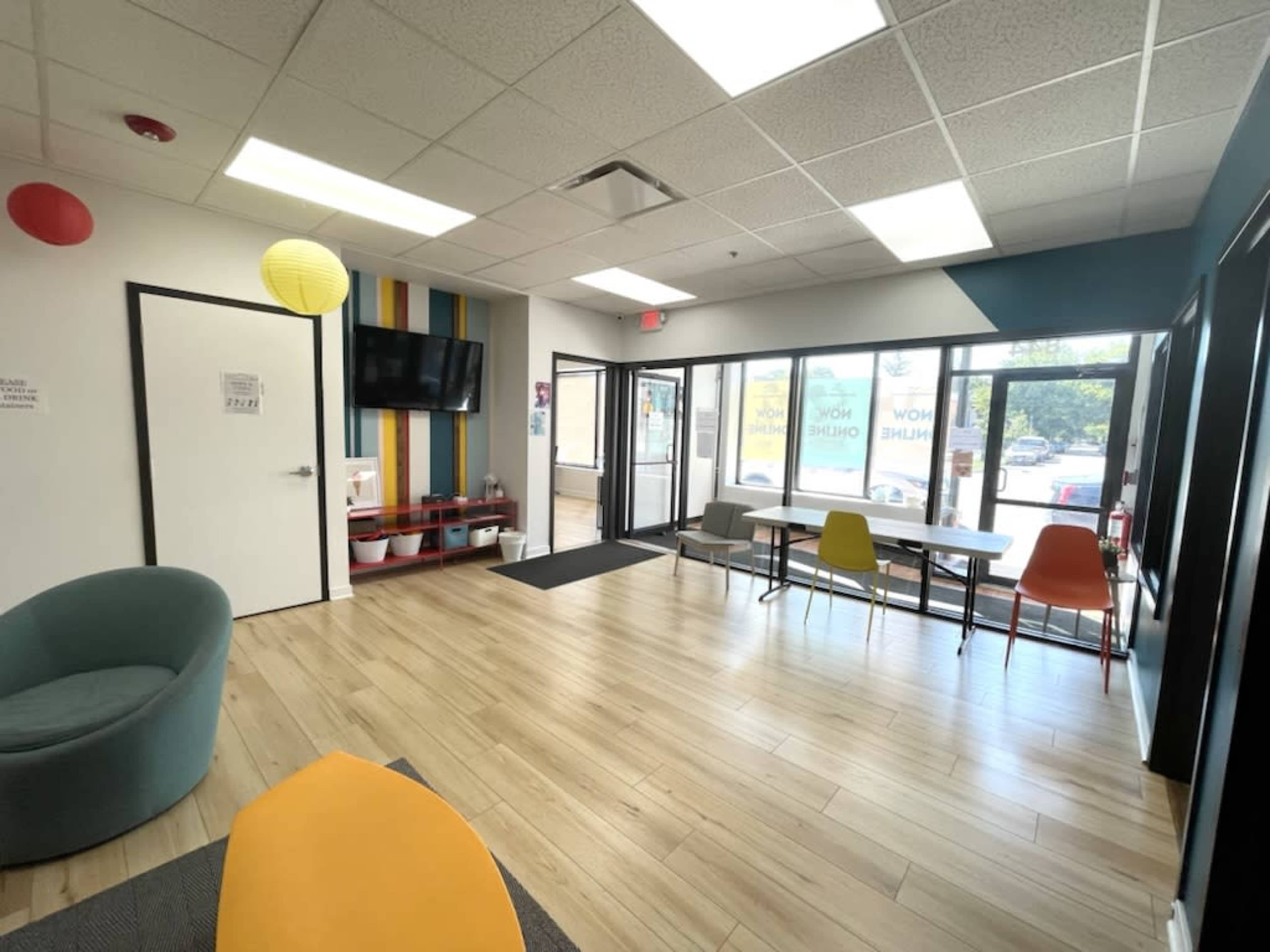 The image shows a bright waiting area with modern furniture, a TV mounted on the wall, and large windows letting in natural light.