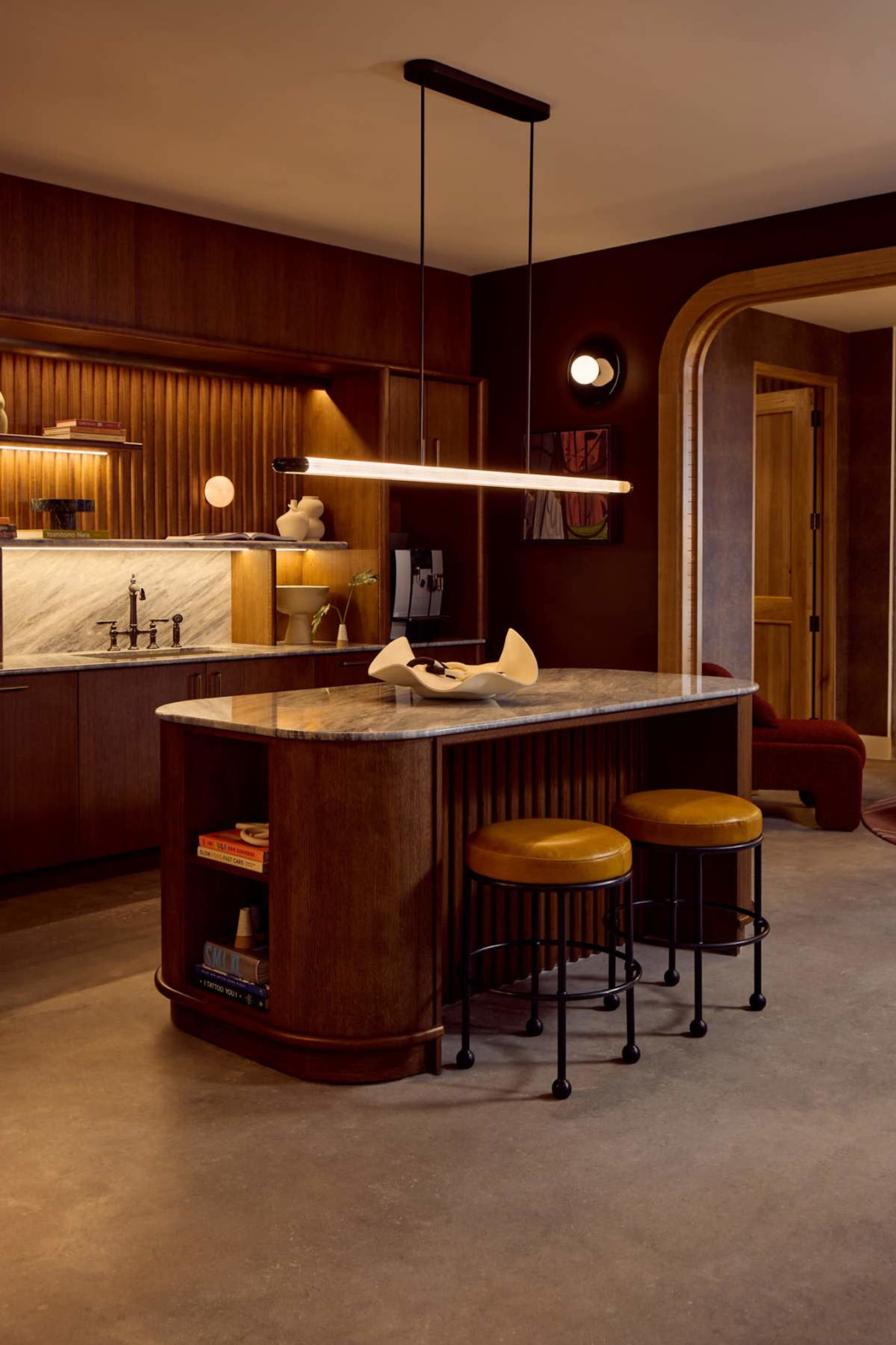 The image features a modern kitchen with a central island, two bar stools, wooden cabinetry, and a marble backsplash.