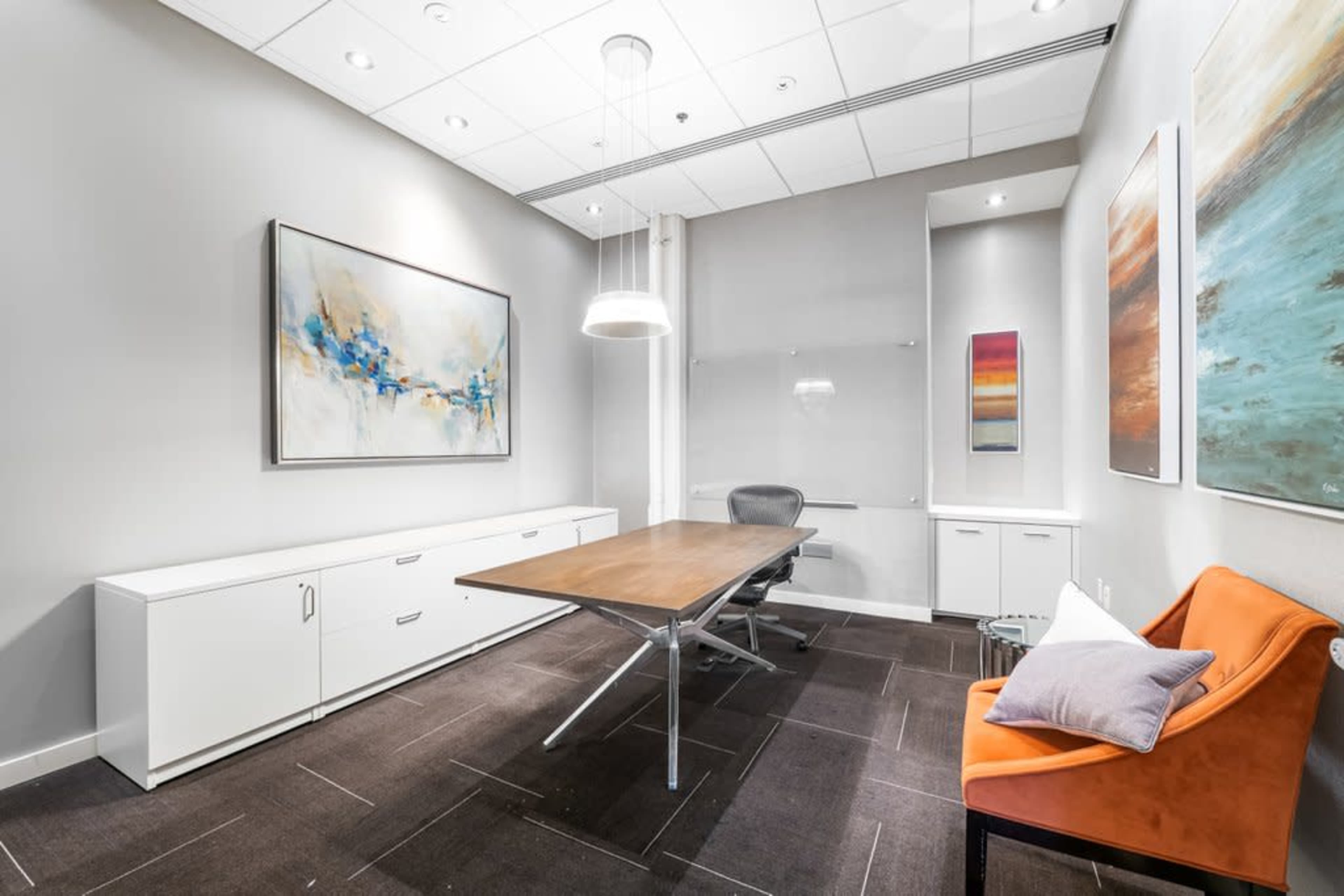The image shows a modern office meeting room featuring a large wooden table, an ergonomic chair, and colorful artwork on the walls.